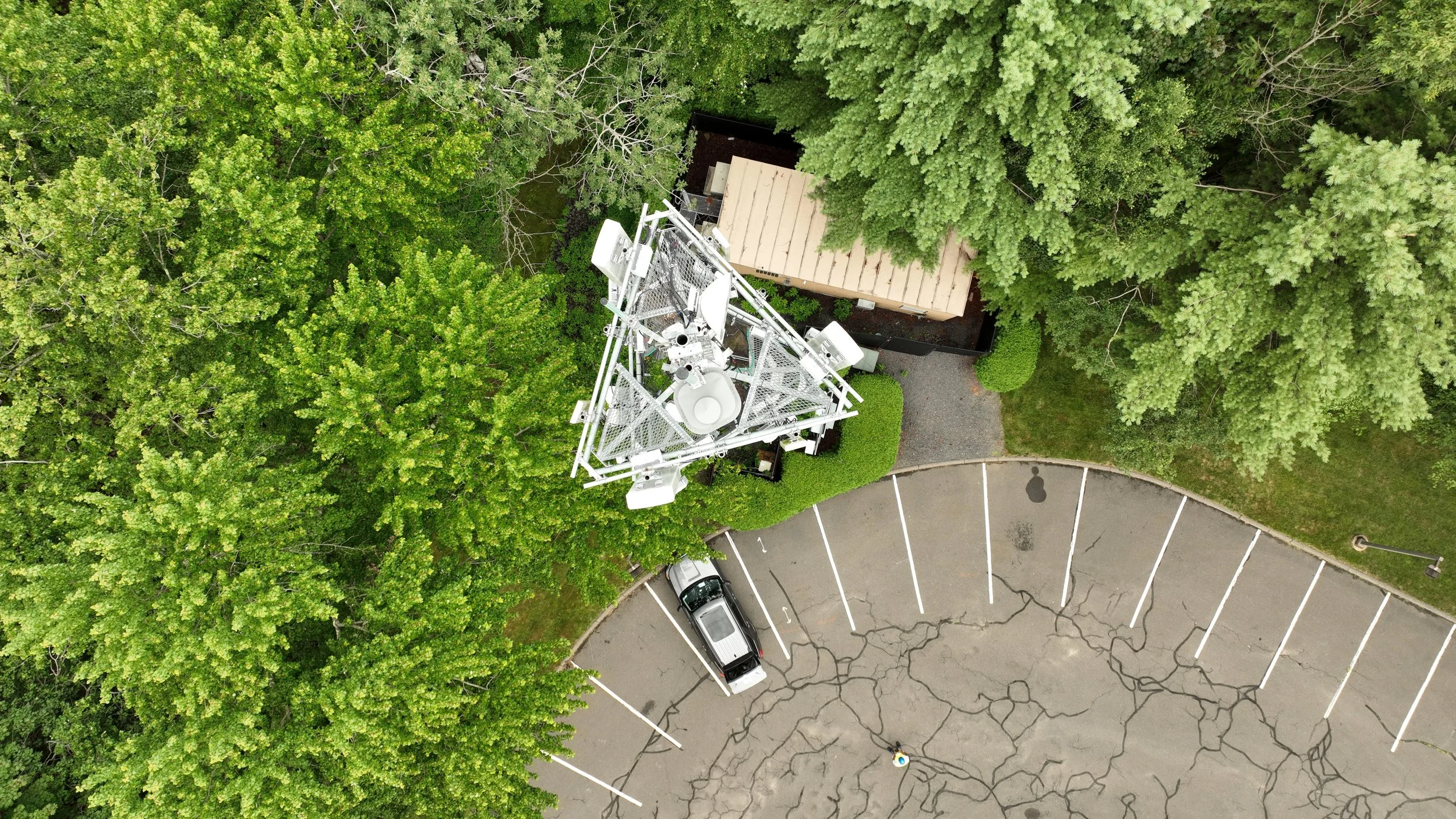 An aerial view of a parking lot with cracked pavement, a single silver car parked near the edge, and a television transmission tower surrounded by green trees.