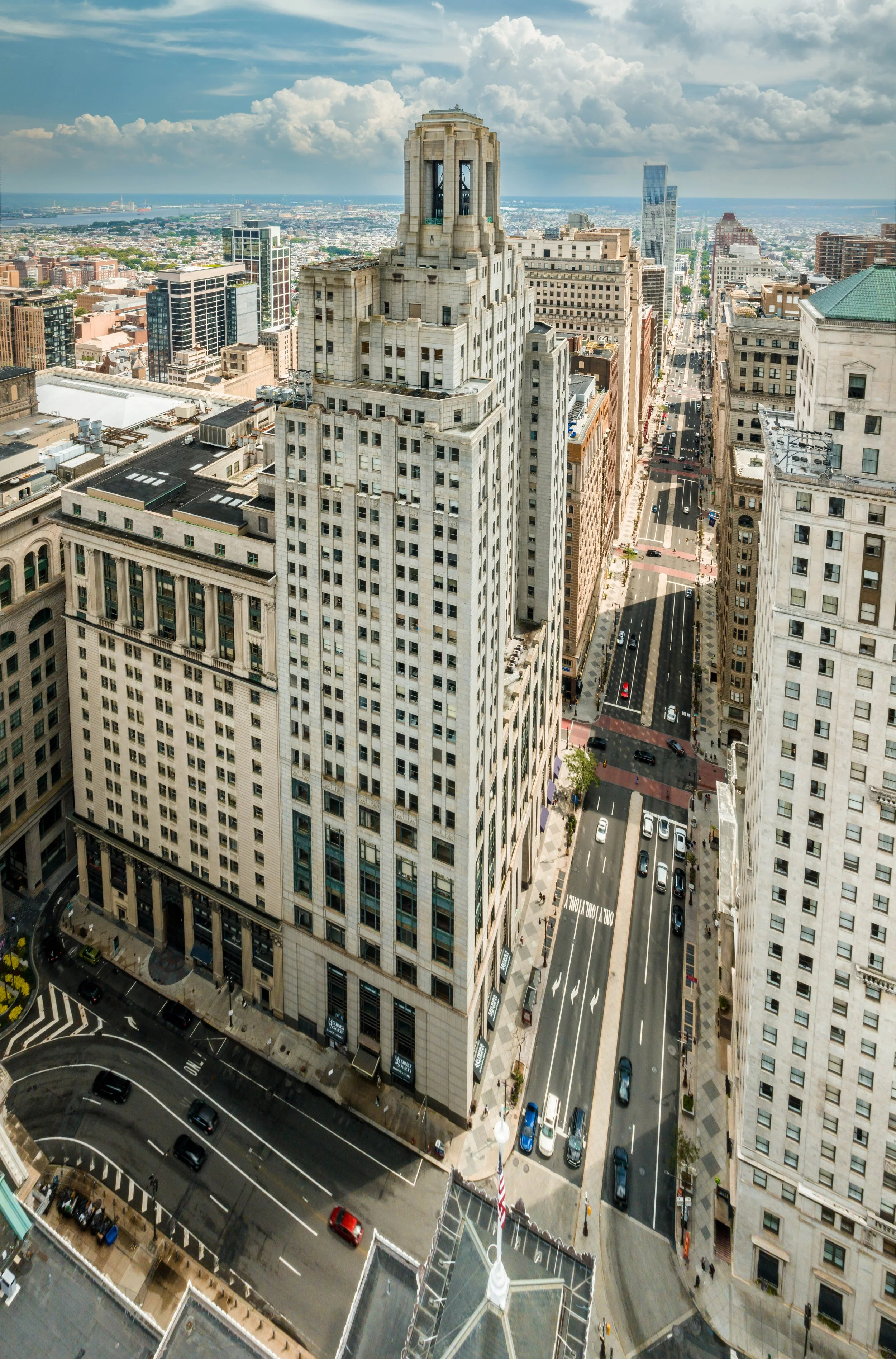 High-angle cityscape view of tall buildings and streets in downtown, with a prominent historic skyscraper in the foreground and modern skyscrapers in the background under a partly cloudy sky.