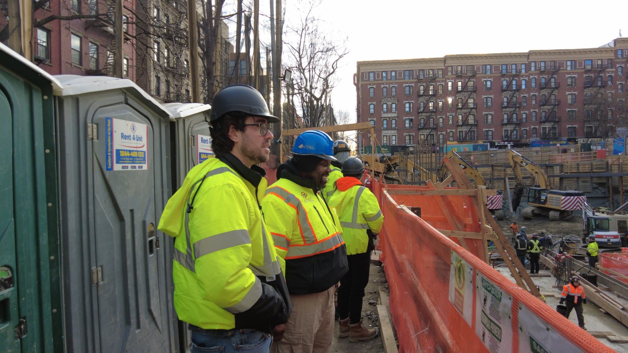 Herman from Those Guys Aerial consulting with Tim, the project manager from SD Builders, in safety gear, observing a construction site with heavy machinery, orange fencing, and a building in the background.