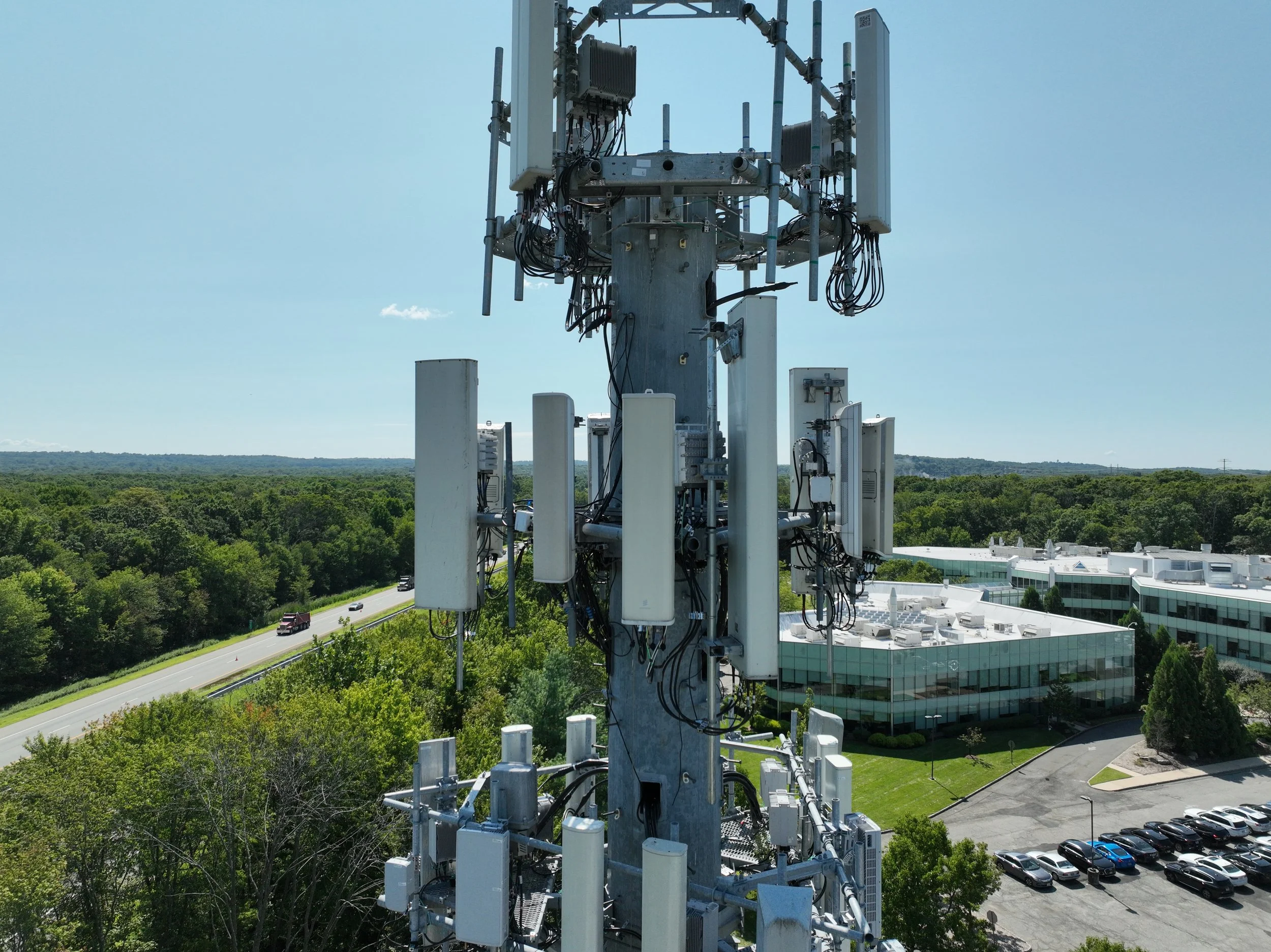 Telecommunications tower with multiple antennas and equipment, surrounded by trees, a parking lot with cars, and office buildings under a clear sky.