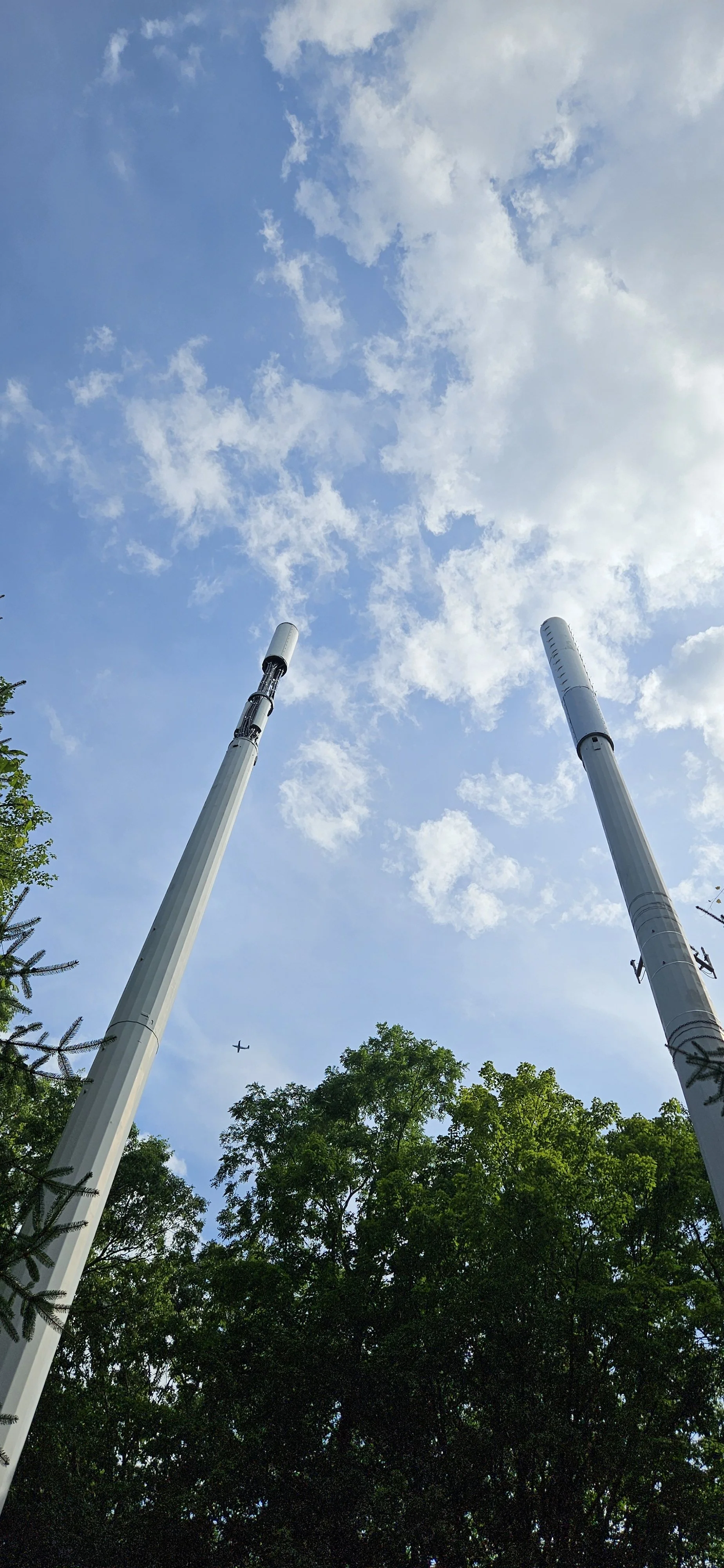 A low-angle view of two tall smokestacks against a blue sky with scattered clouds, surrounded by green trees and an airplane flying in the distance.