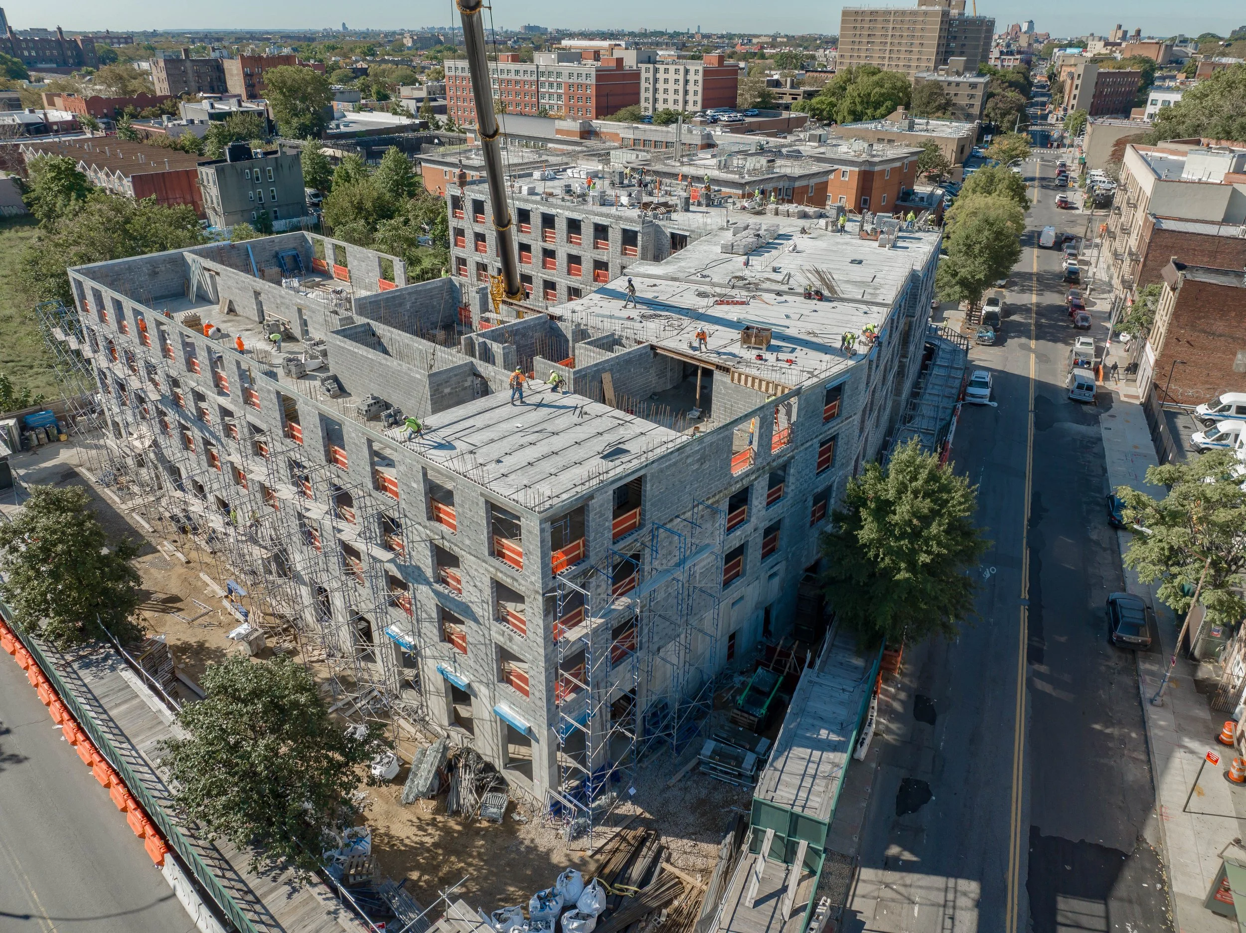 An aerial view of a multi-story building under construction in an urban area, with scaffolding around the structure, construction workers on the roof, and a crane in operation, surrounded by streets and other buildings.