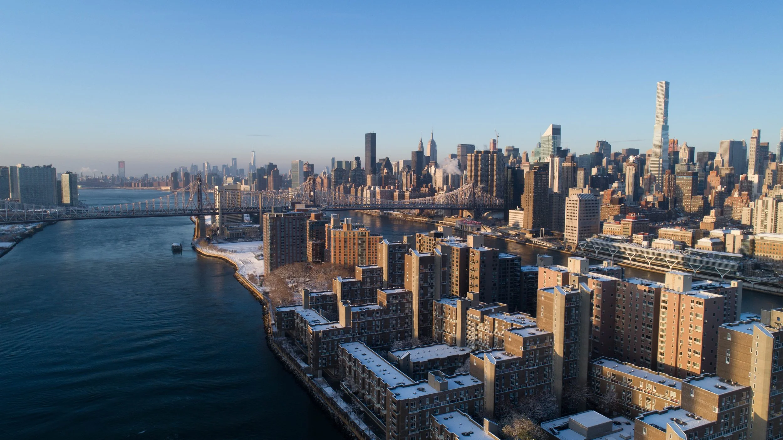 Aerial view of New York City skyline with bridges over the East River, tall skyscrapers, and a clear blue sky on a cold winter day.