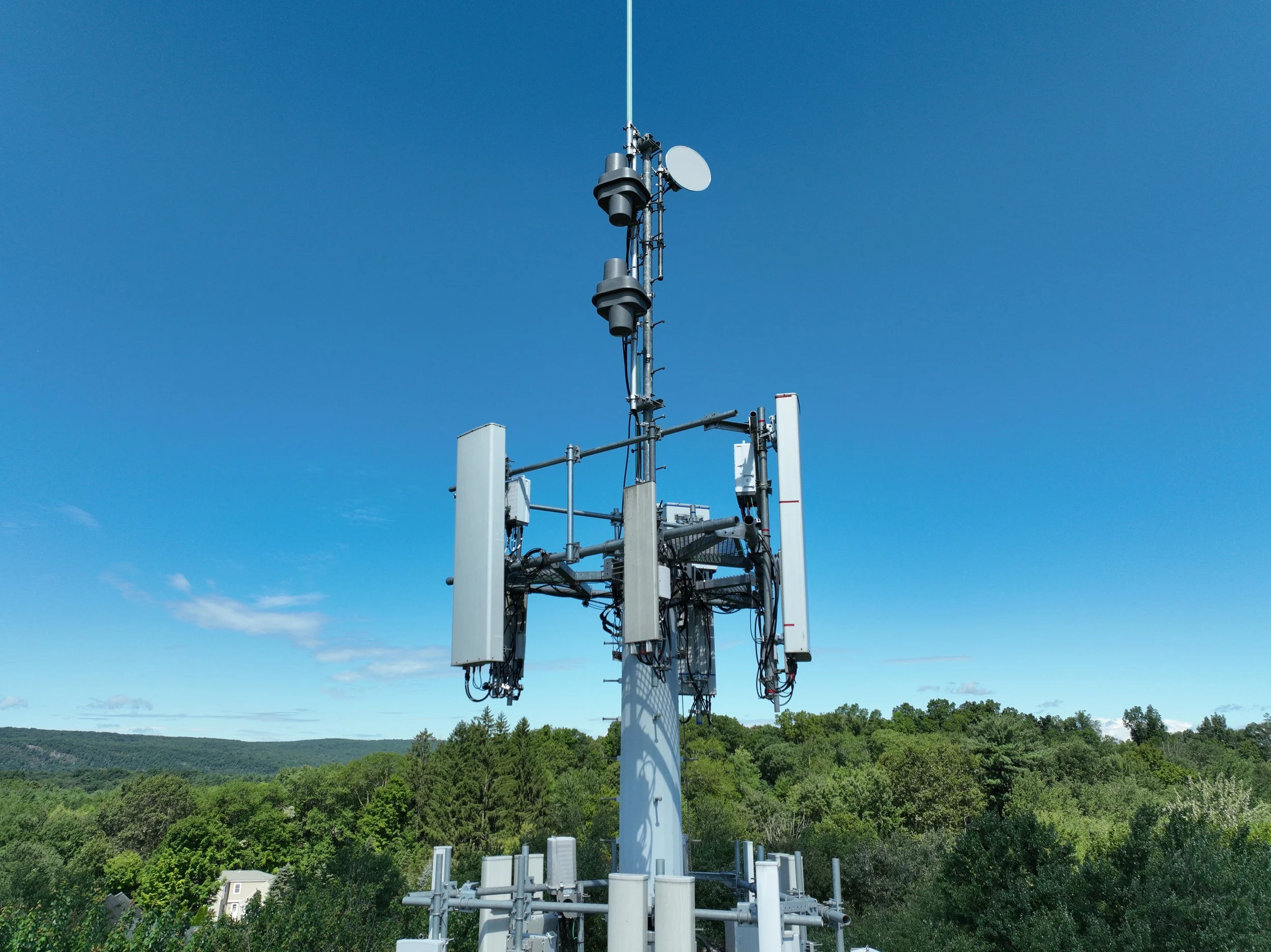 Cell tower with antennas and equipment, set against a bright blue sky and green landscape.