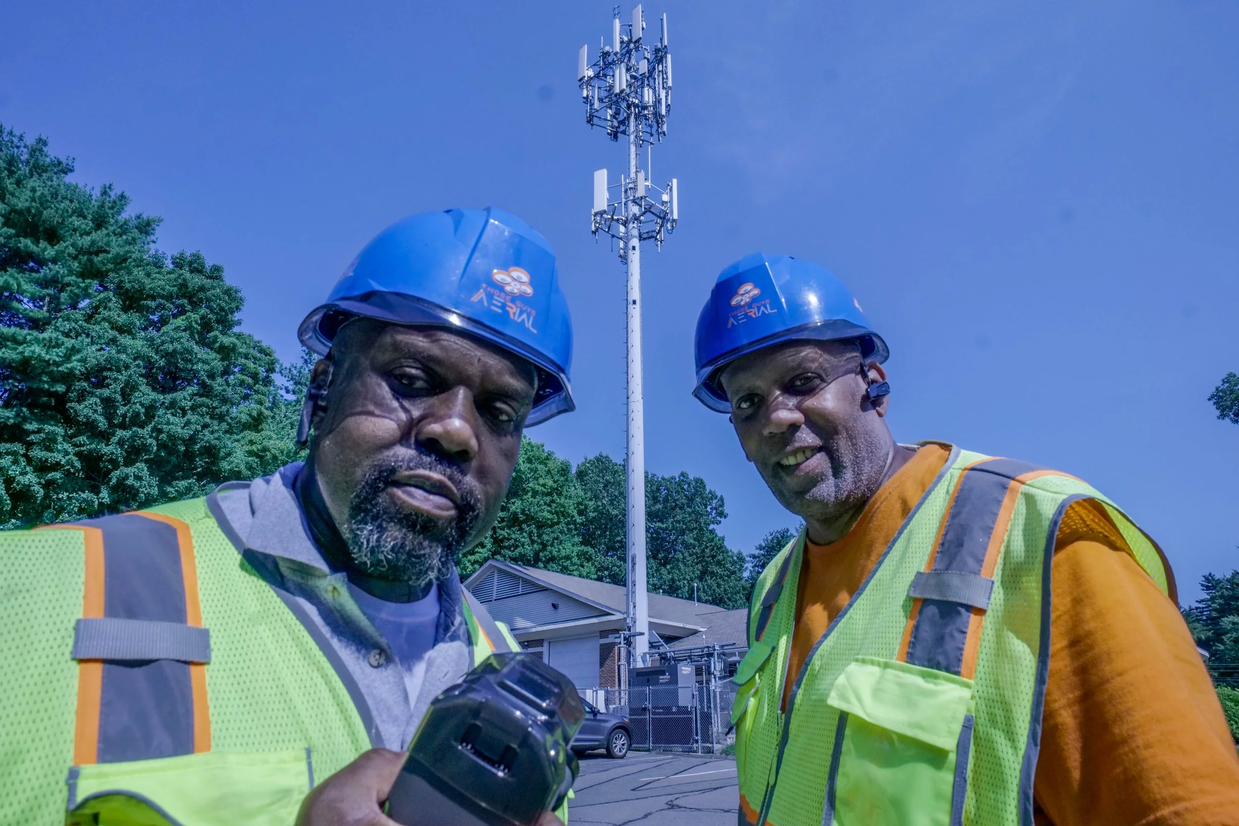 Herman and Lawrence are standing outside near a tall cell tower, doing inspections