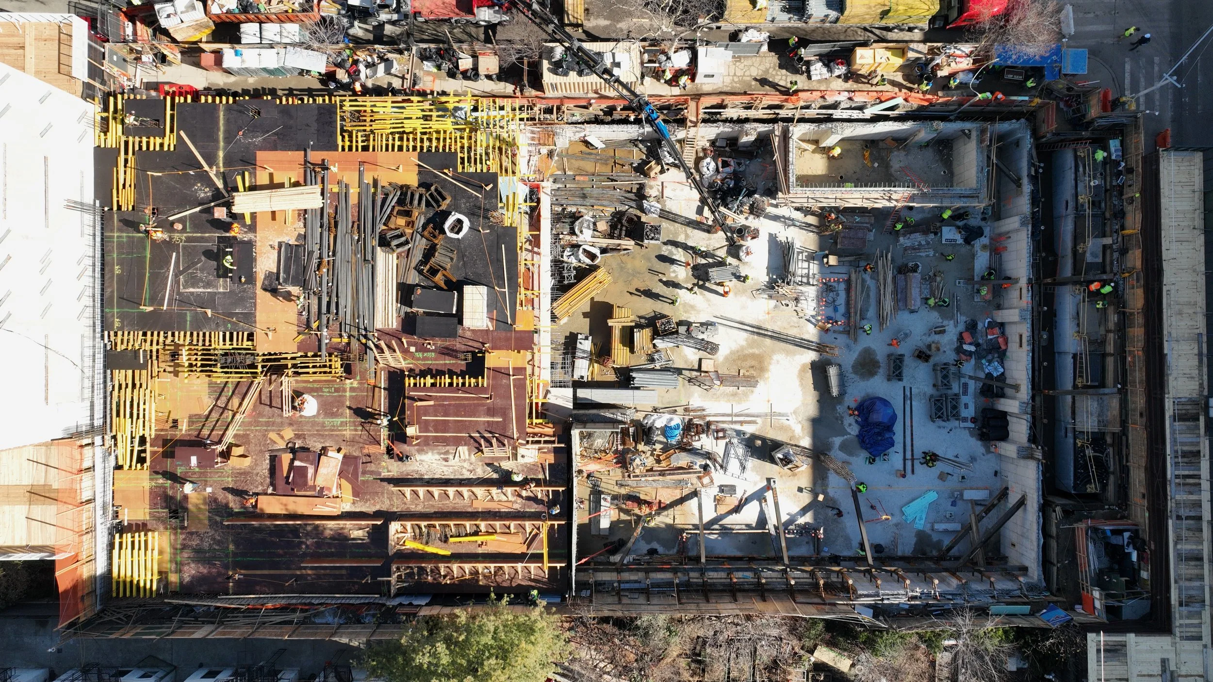 An aerial view of a construction site divided into two sections. The left section shows the early framing phase with timber studs. The right section has concrete floor with construction workers and equipment assembling structures.
