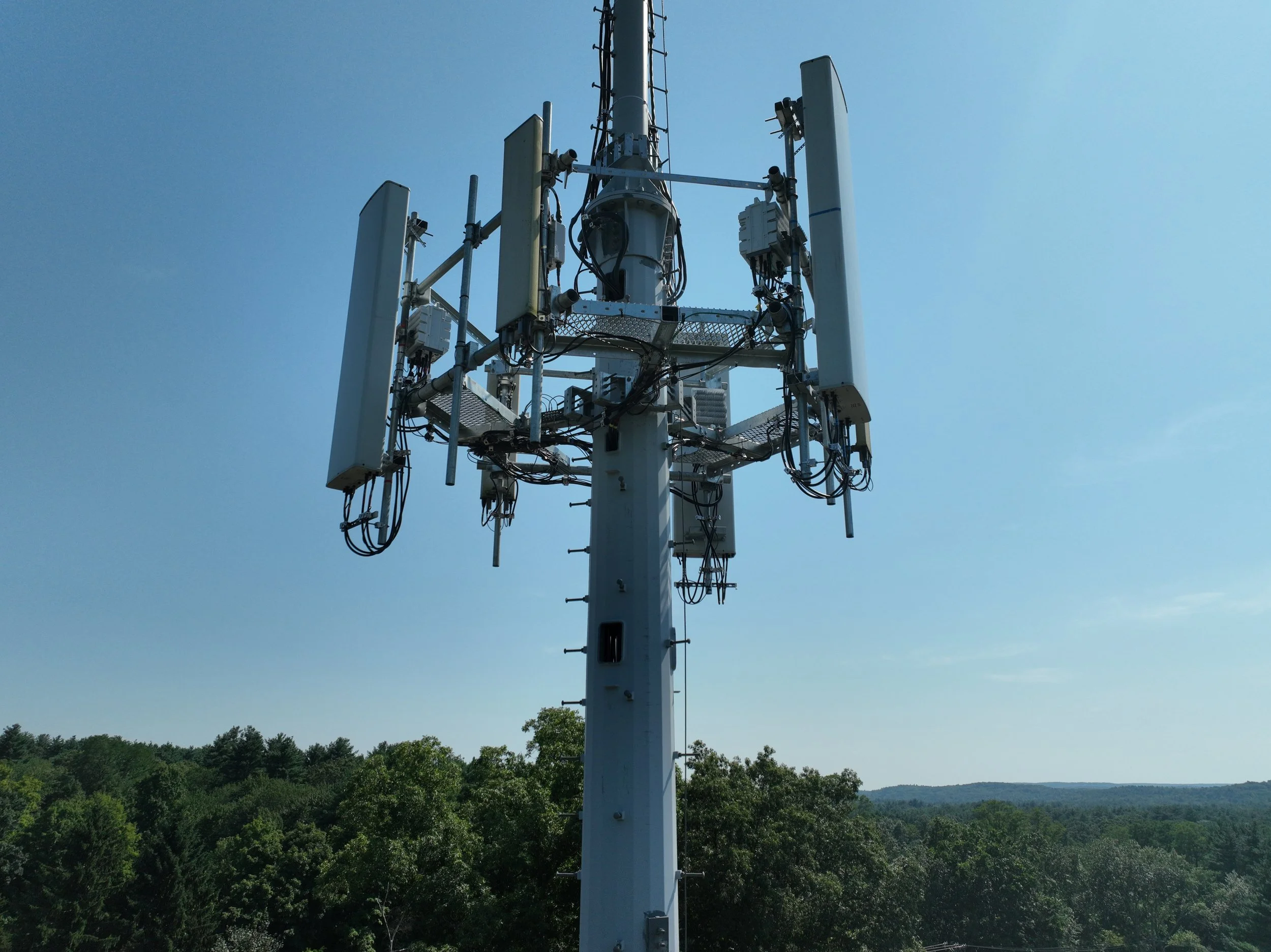 A tall telecommunications tower with multiple antennas and electronic equipment against a blue sky, surrounded by green trees and hills in the background.