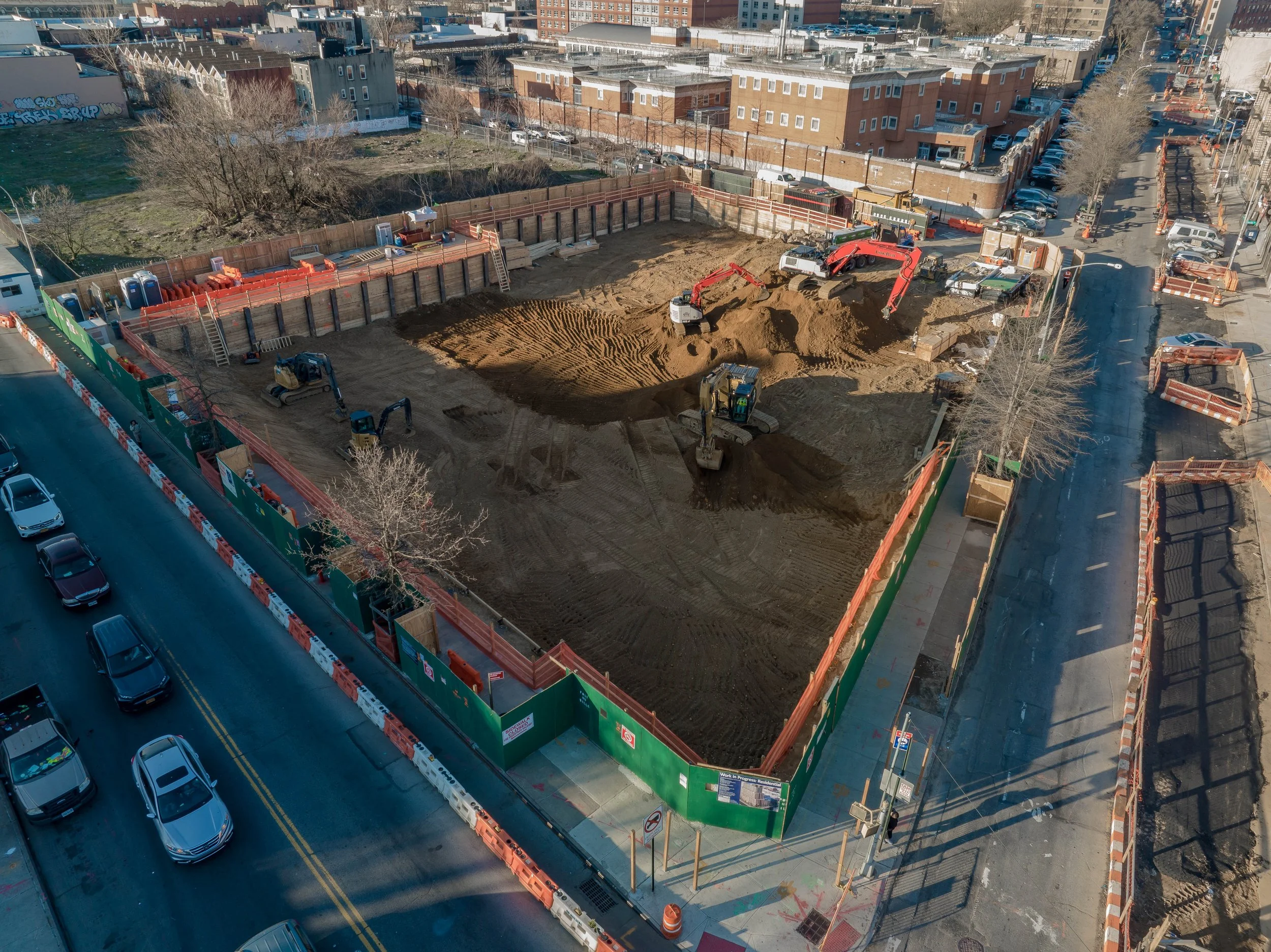 Construction site in an urban area with several heavy machinery excavating and moving dirt. The site is enclosed with orange and green safety barriers, and there are ladders and portable toilets on the perimeter. Adjacent roads with parked and moving cars surround the site.