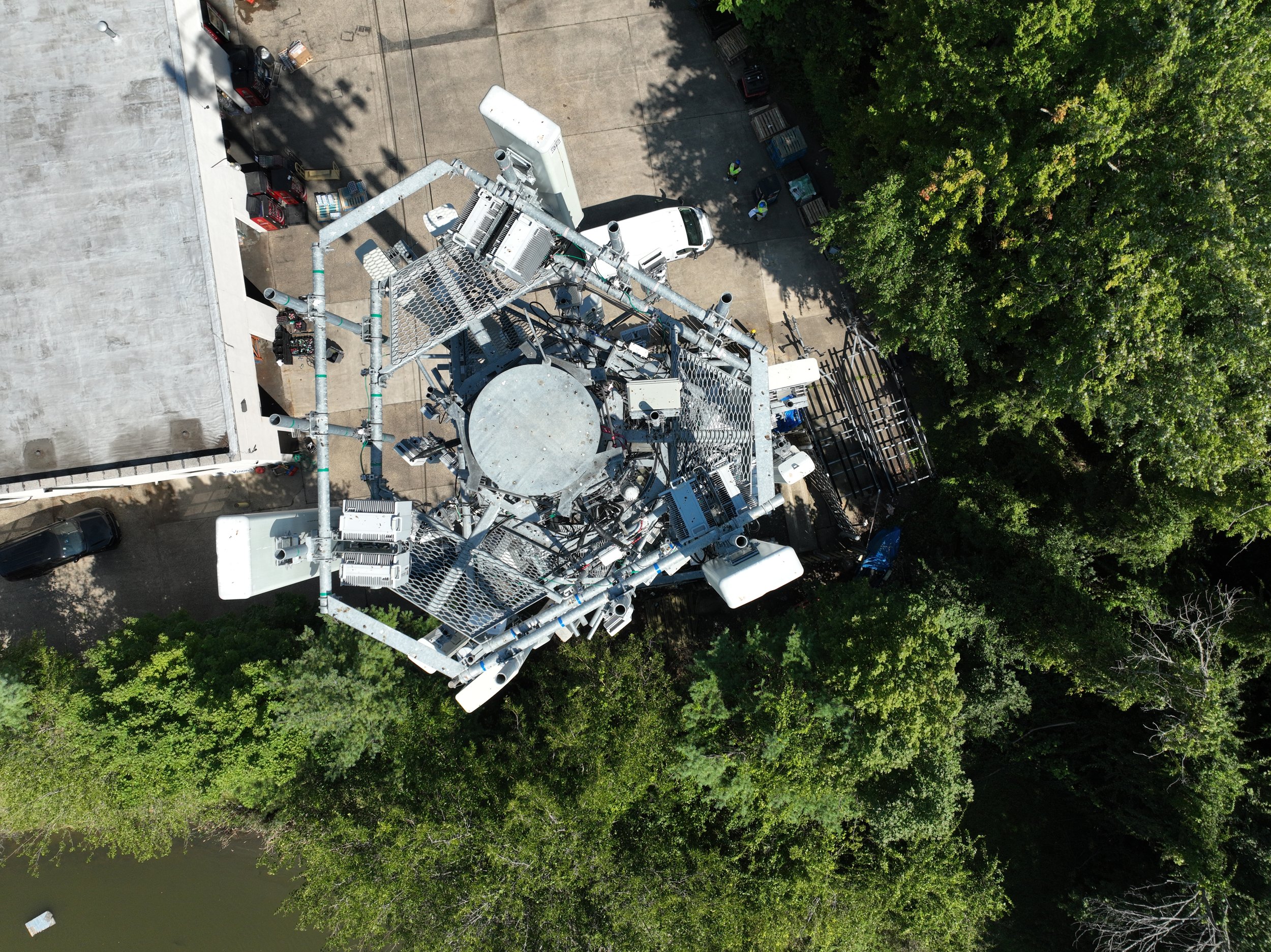 An aerial view of a cell tower surrounded by trees, parked vehicles, and a building.