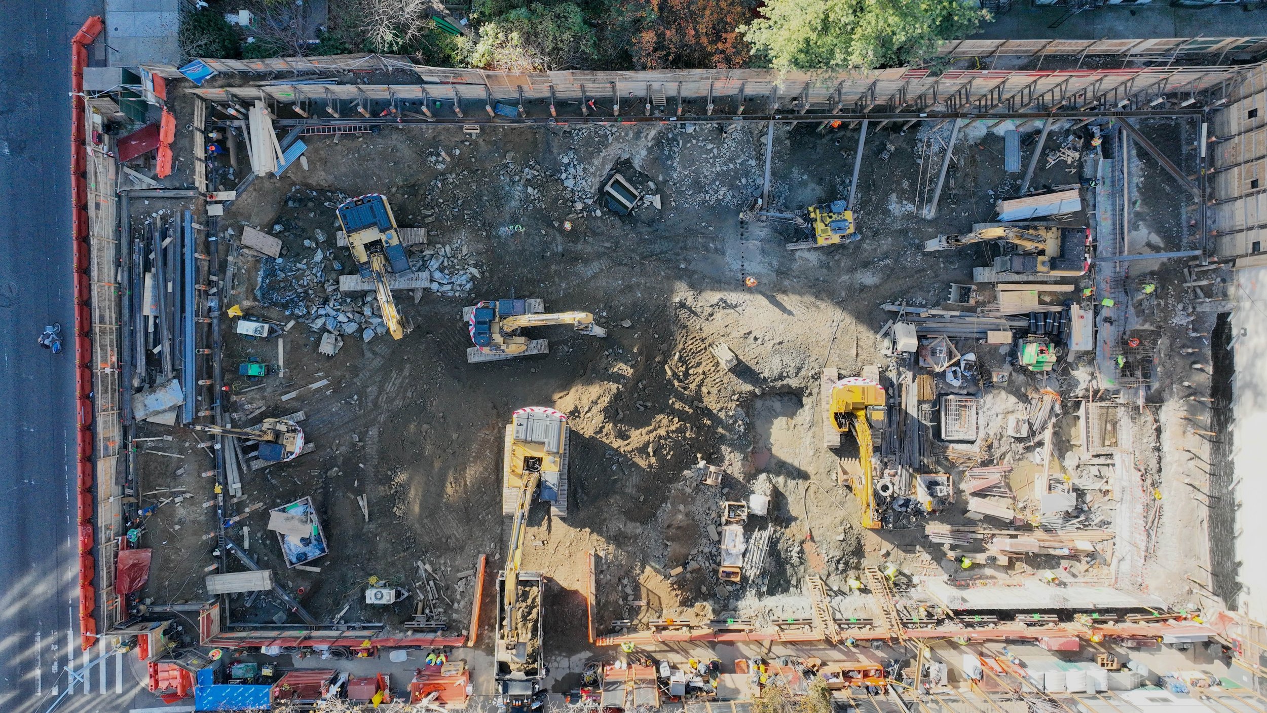 An aerial view of a construction site with several excavators and construction workers working on a building foundation, surrounded by safety barriers and city street.