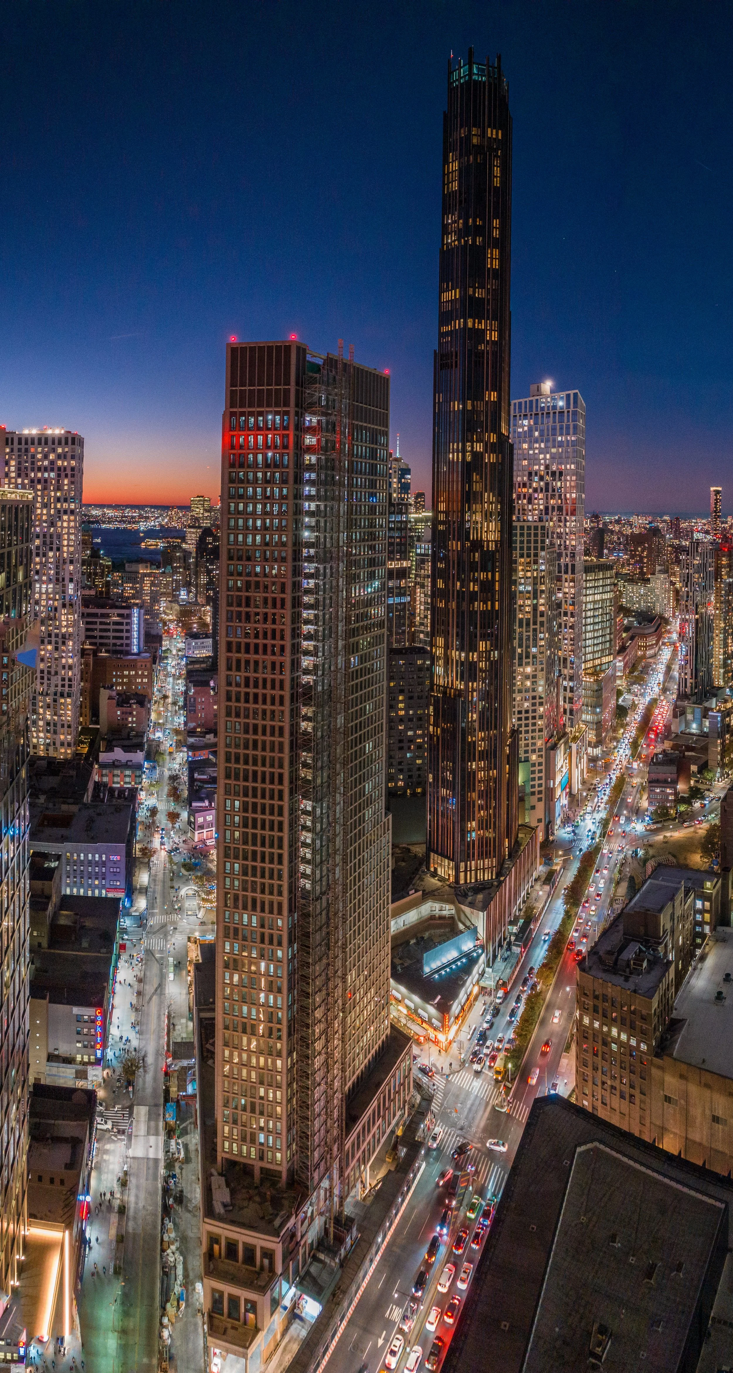 Nighttime cityscape with tall illuminated skyscrapers, busy streets with car headlights, and a dark blue sky with a hint of sunset on the horizon.