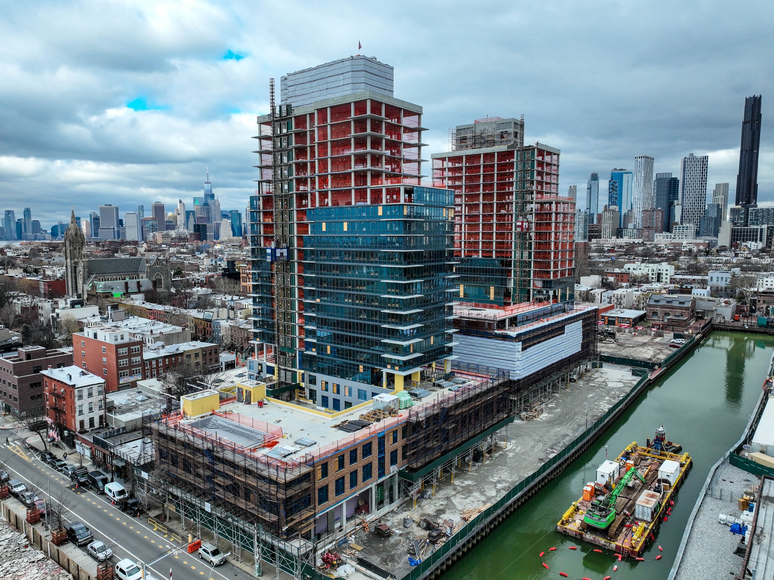 Aerial view from Gowanus Canal with two high-rise buildings under construction, surrounded by other buildings, with a cloudy sky in the background.