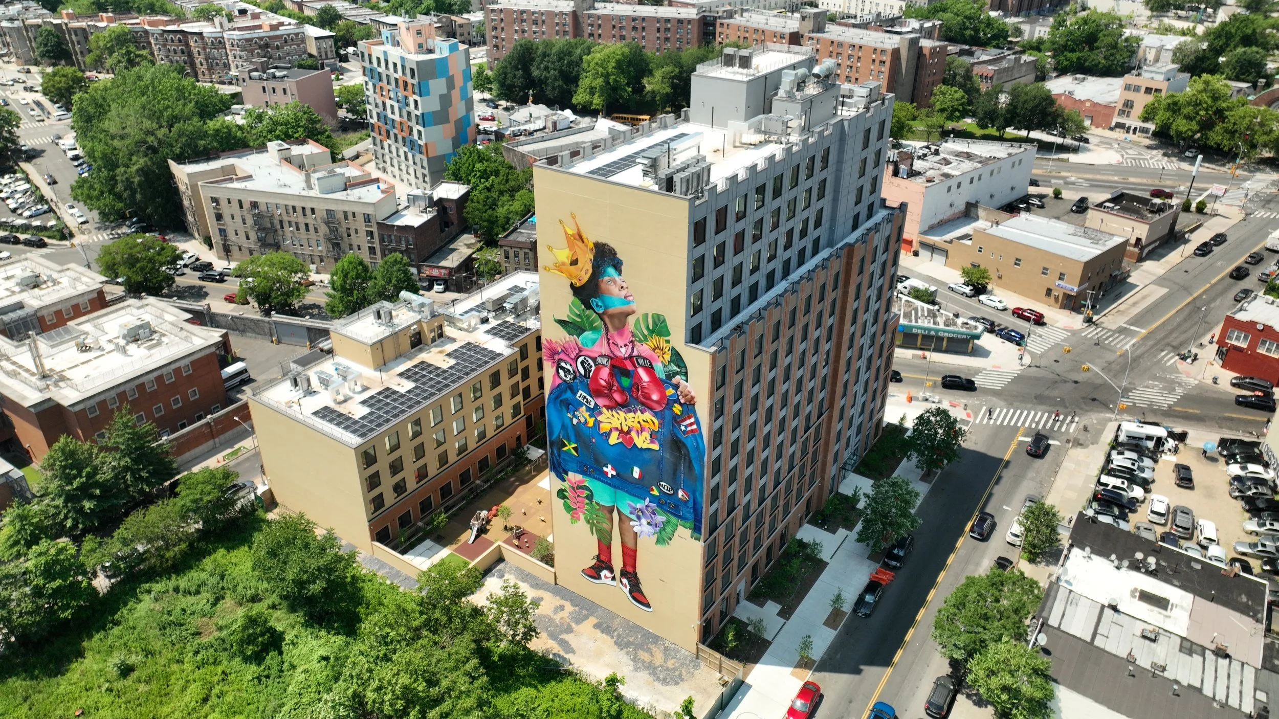 Large colorful mural of a woman with a crown and boxing gloves on the side of a tall building in an urban neighborhood, surrounded by trees and other buildings.