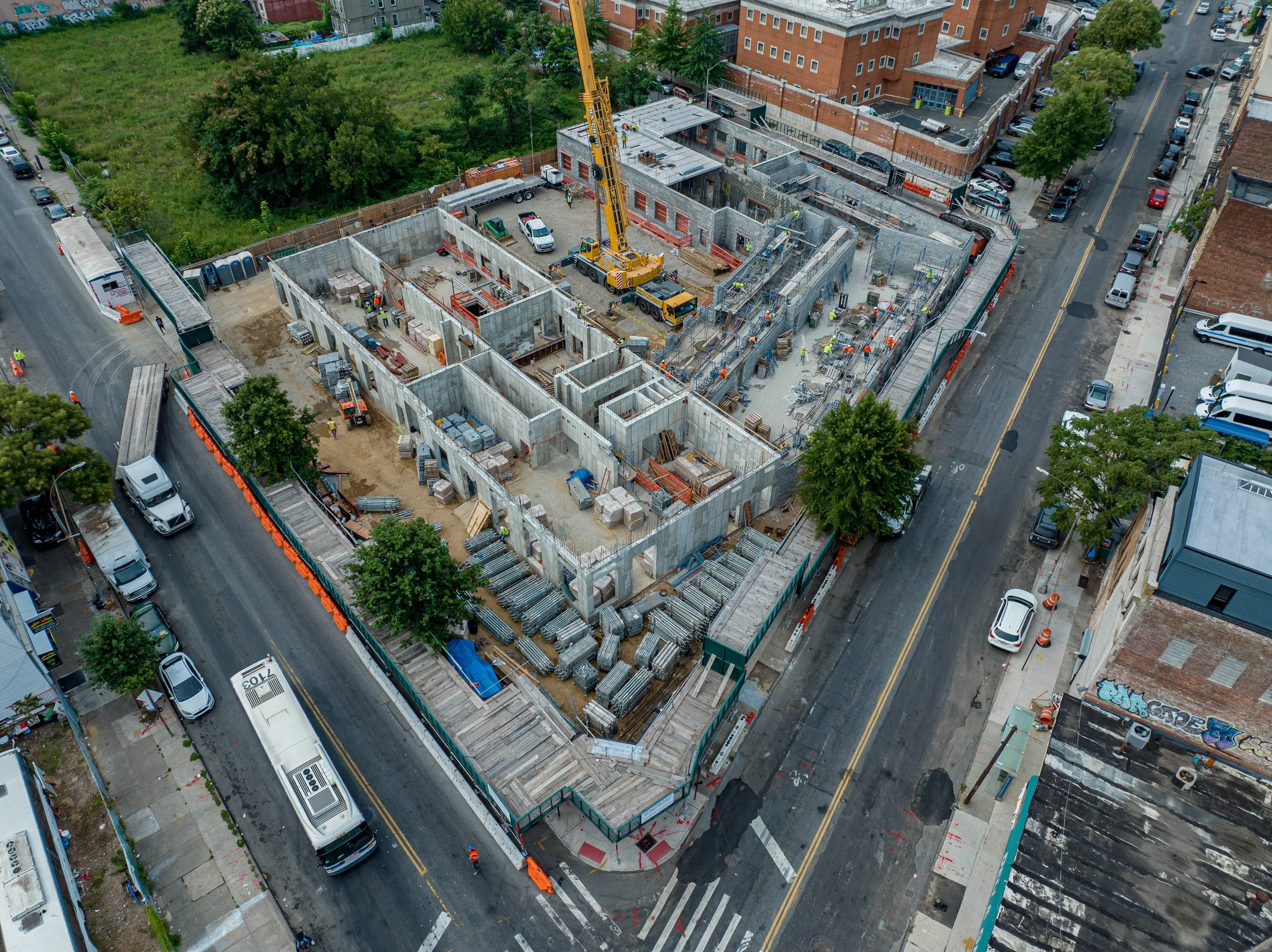Aerial view of a construction site on a city street, with concrete building framework and workers, surrounded by parked cars and adjacent buildings.