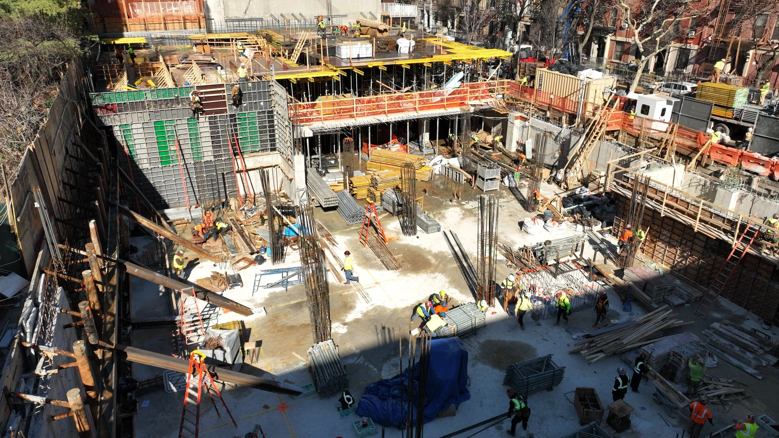 Construction site with workers building a multi-level structure, using steel rebar, wooden formwork, and cranes, in an urban area with trees and buildings nearby.