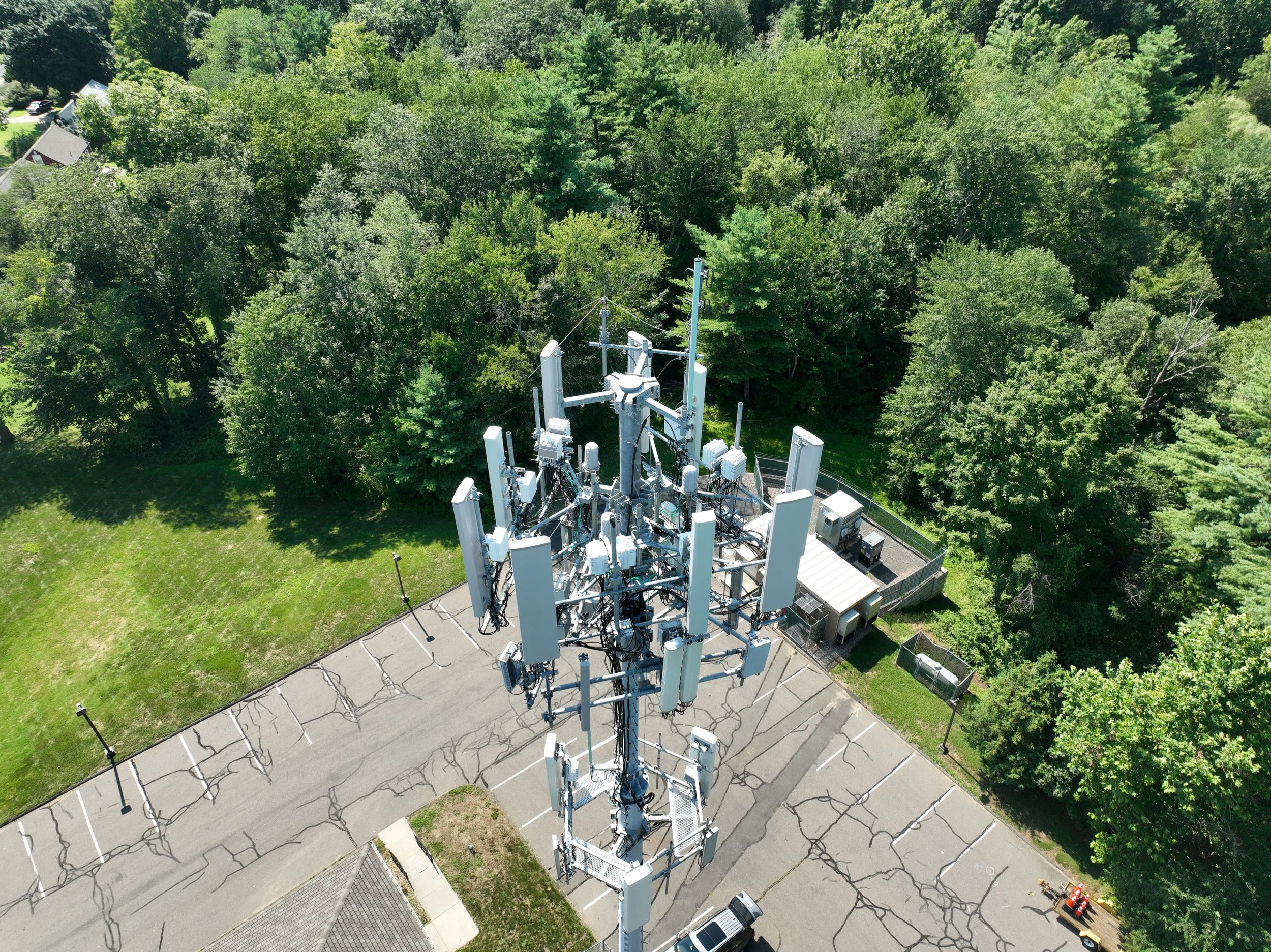 An aerial view of a cell tower with multiple antennas surrounded by trees and a parking lot.