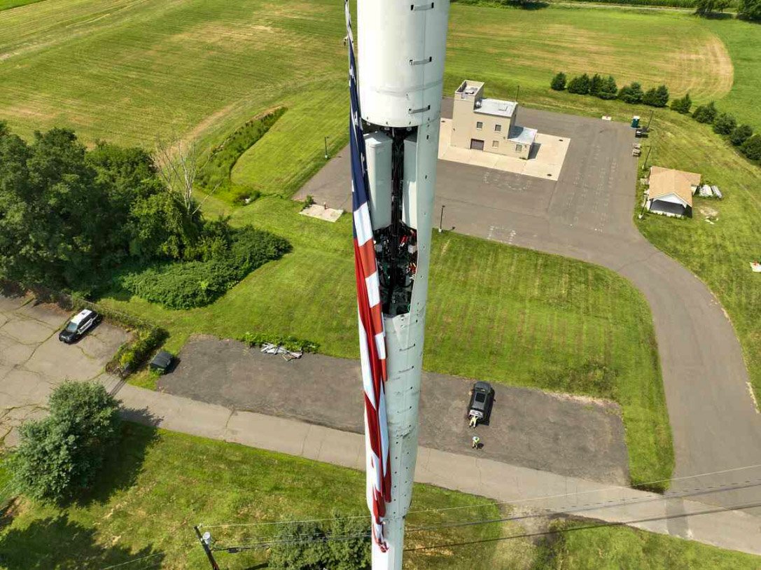 A large radio or cell tower with a partially disassembled American flag hanging down the side, taken from above on a sunny day in a suburban area with green lawns, trees, and buildings.
