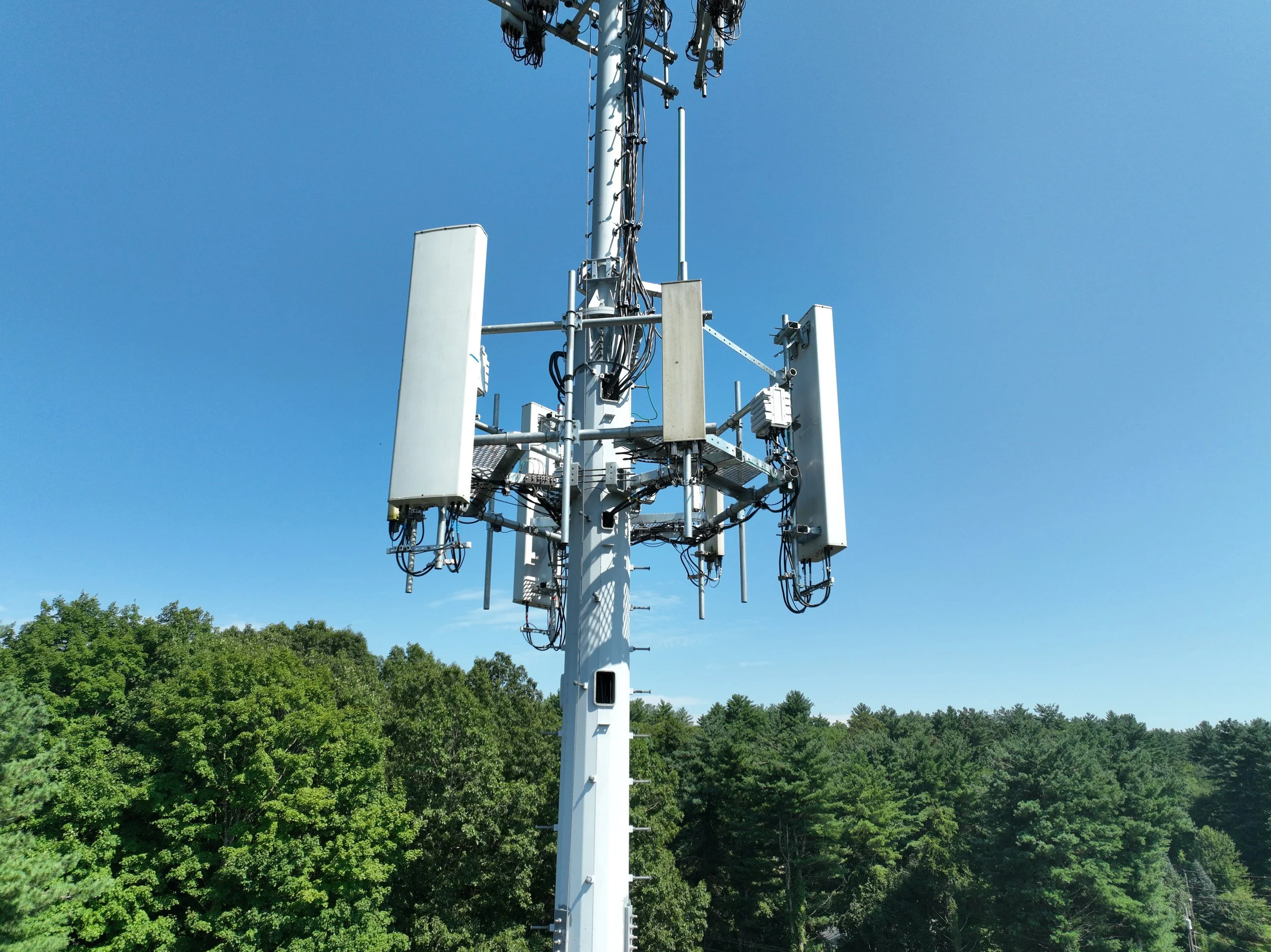 Tall cellular tower with multiple antennas and equipment mounted, against a background of green trees and a clear blue sky.