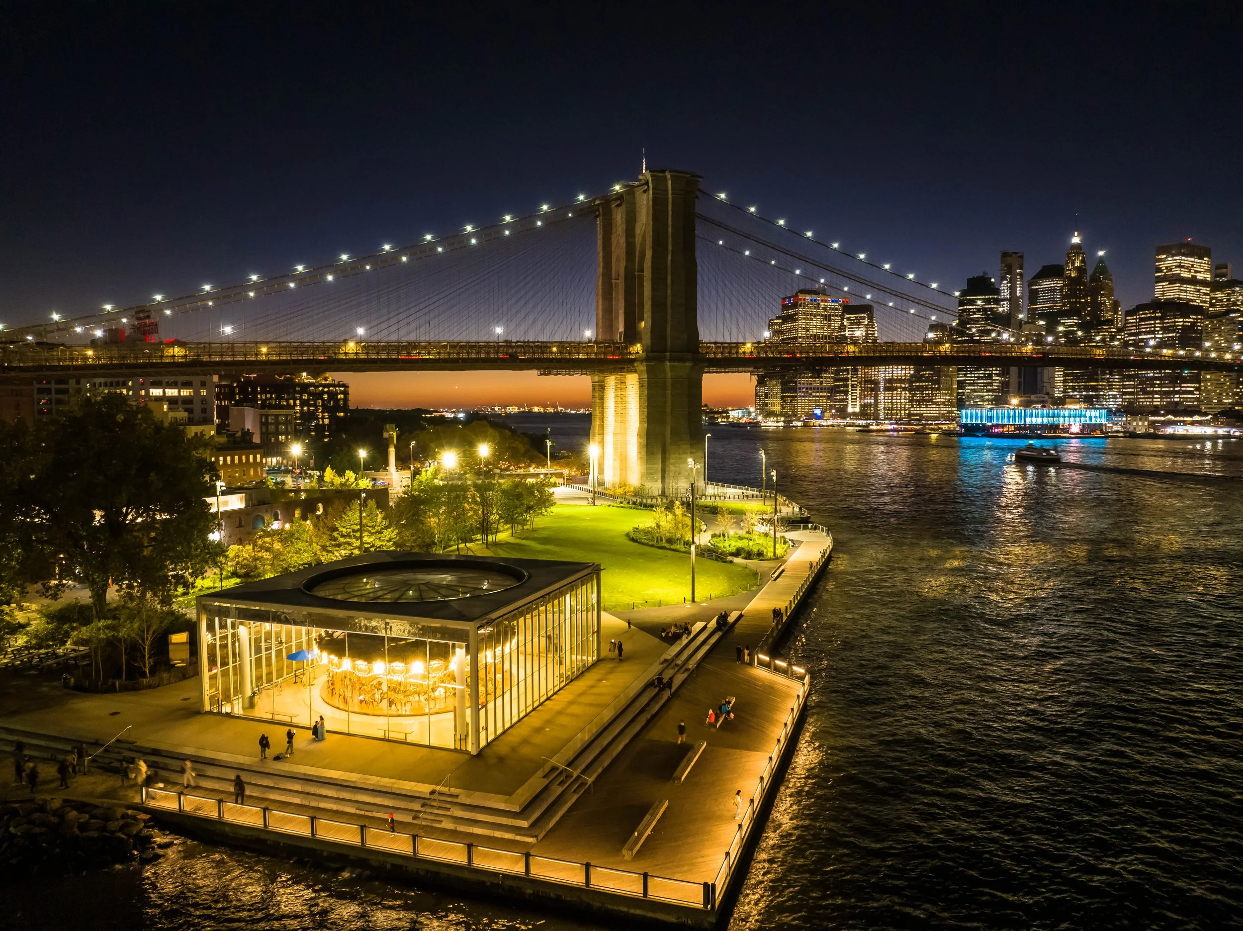 Night view of Brooklyn Bridge with illuminated city skyline behind, and a glass carousel pavilion on a waterfront park in New York City.