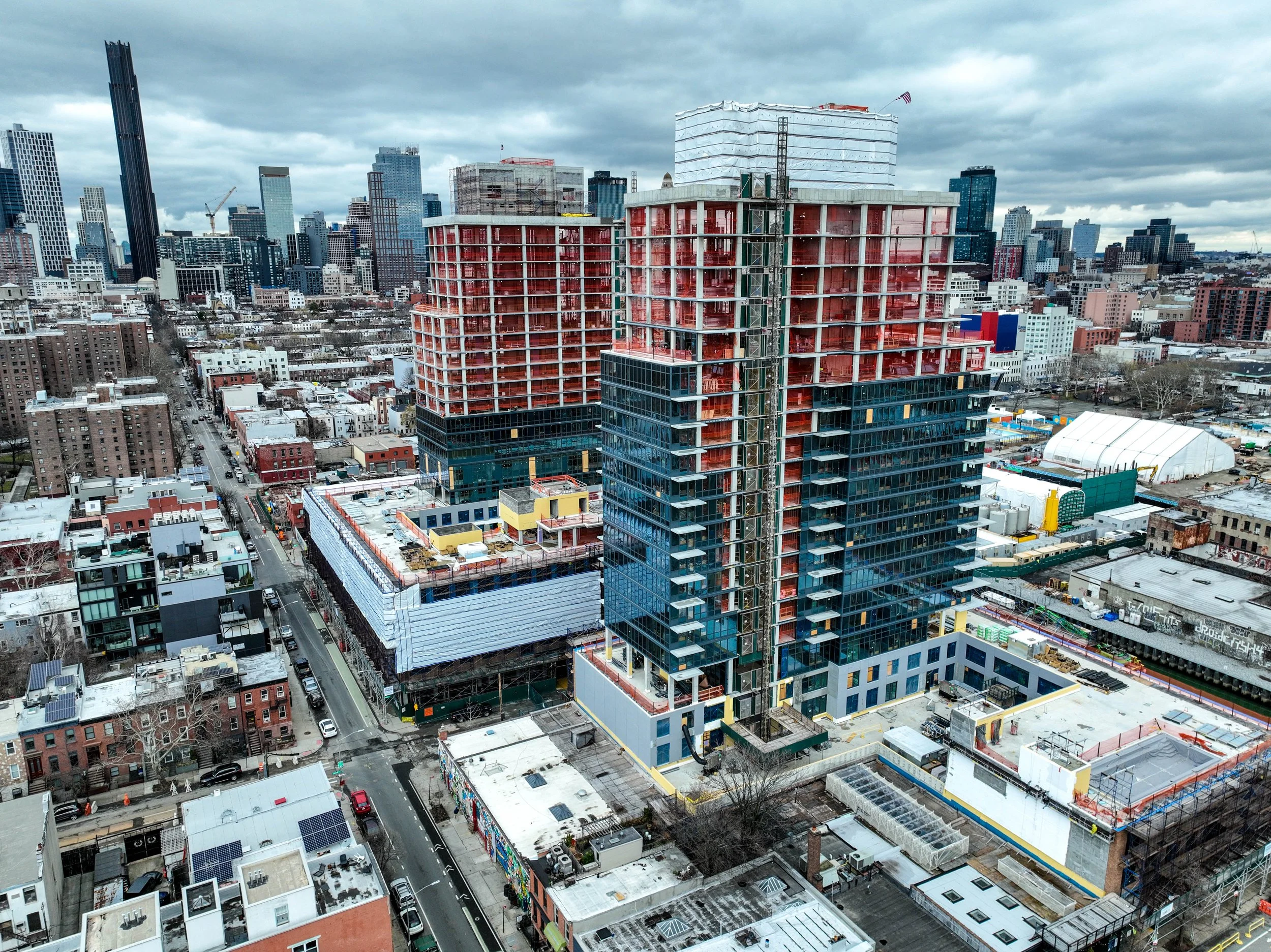 Aerial view from Gowanus Canal with two high-rise buildings under construction, surrounded by other buildings, with a cloudy sky in the background.
