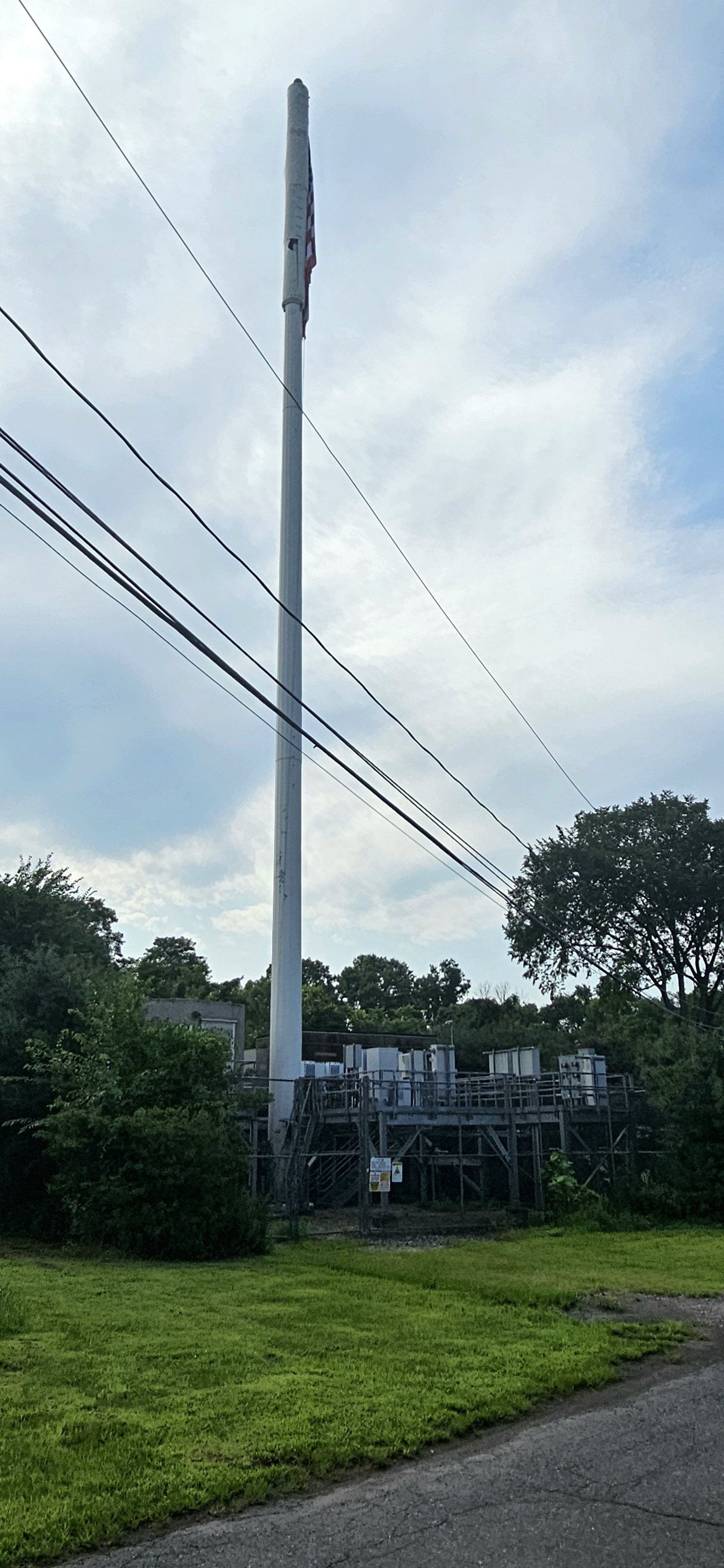 A tall utility pole with wires extending from it, set against a partly cloudy sky, with trees and electrical equipment at the base.
