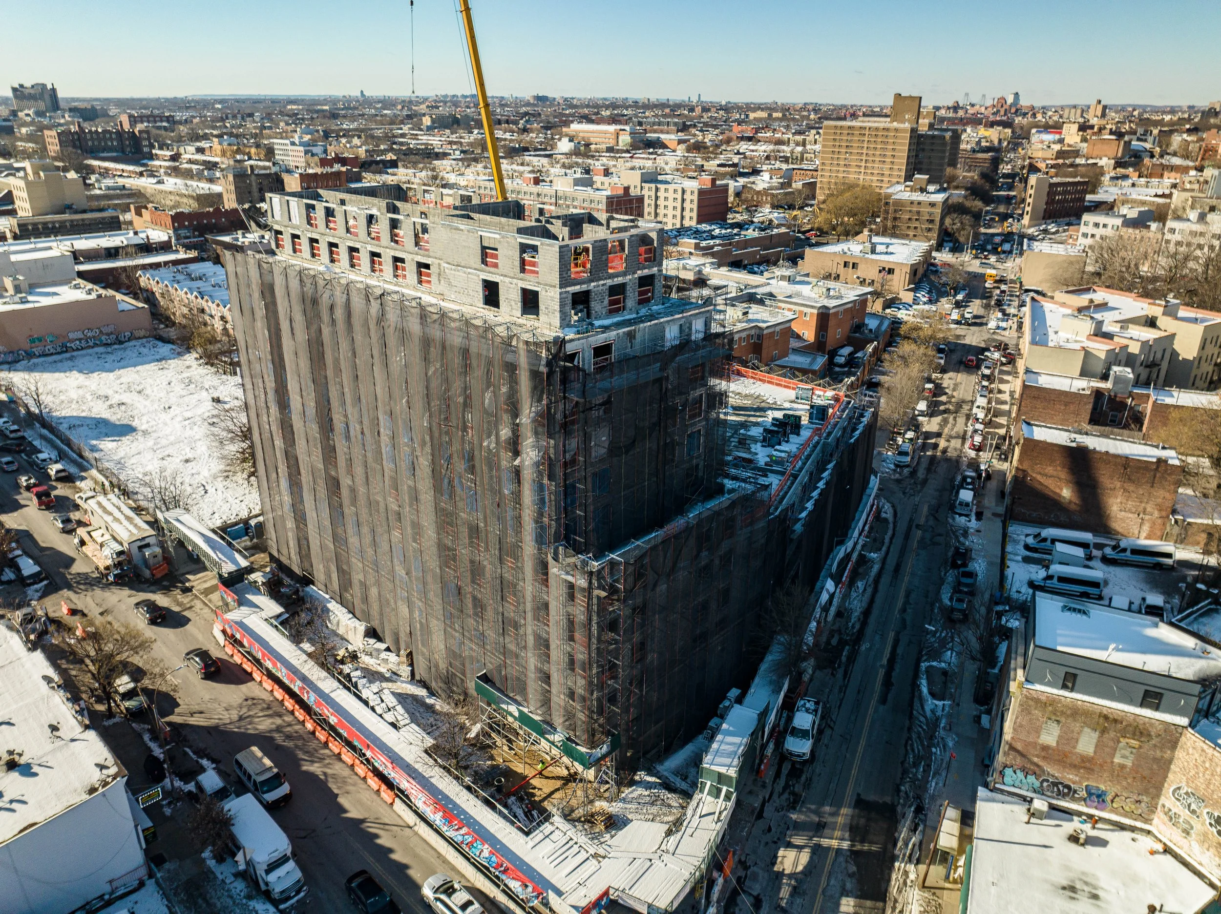 Aerial view of a snowy cityscape showing a building under construction, surrounded by snow-covered roads, parked cars, and other buildings.