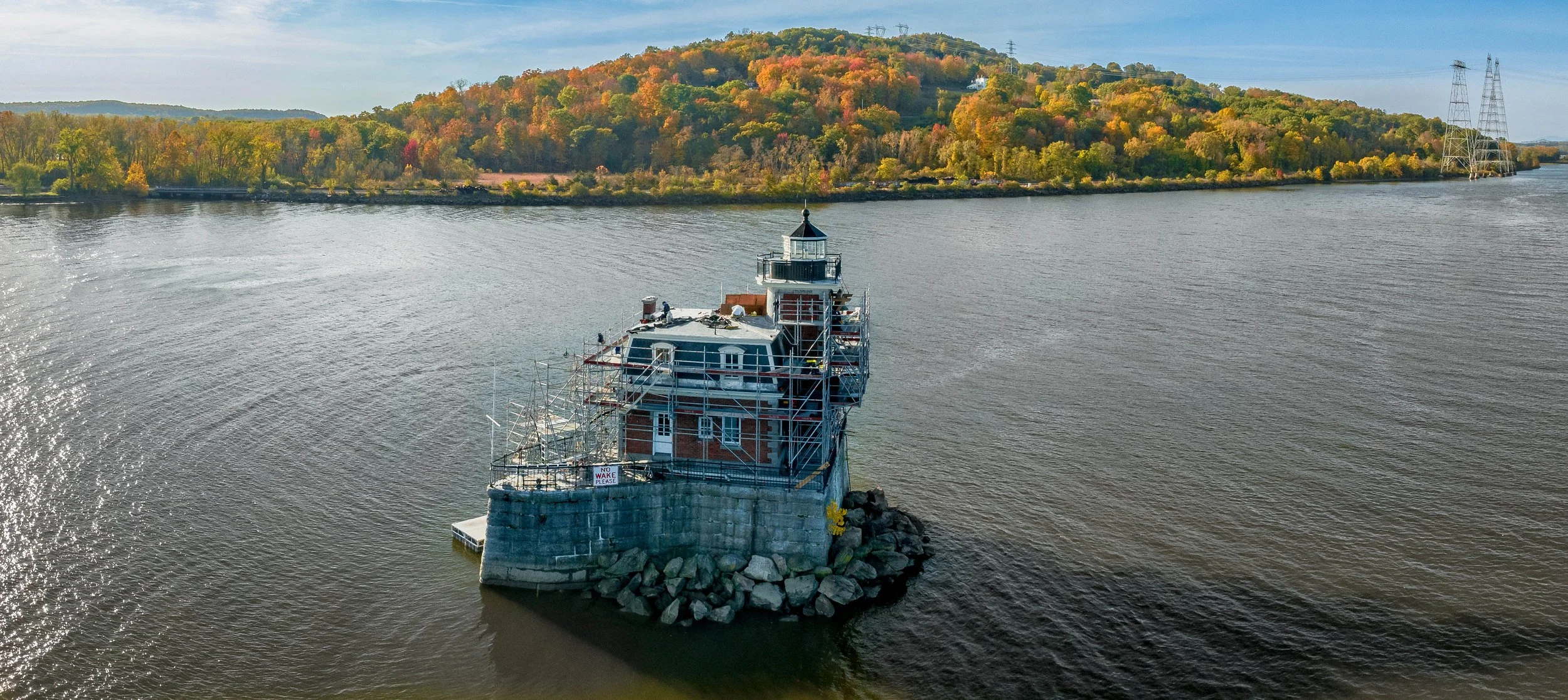 A lighthouse on a small rocky island in a river, with construction scaffolding around it, and a forested hill in the background with autumn-colored trees.
