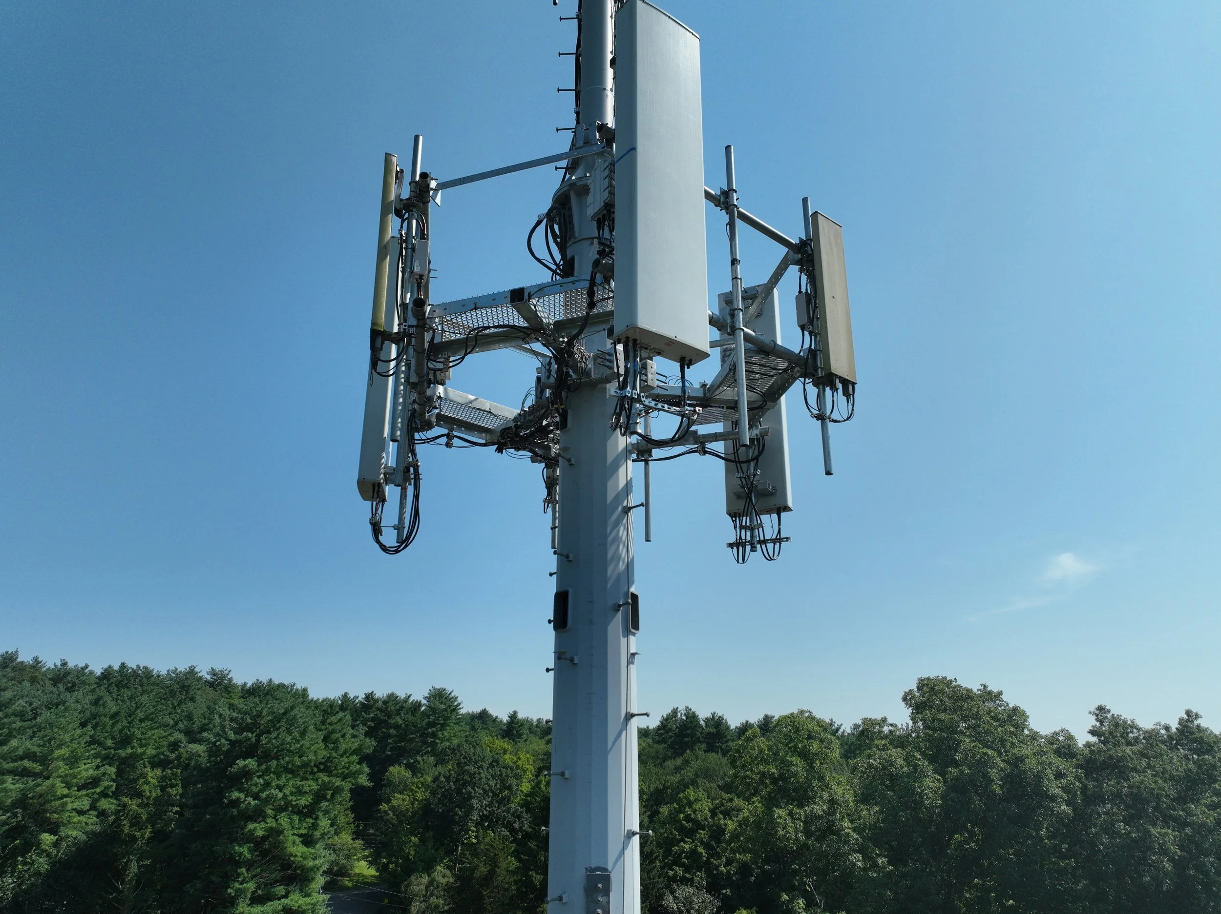 A tall cellular communication tower with multiple antennas and equipment against a clear blue sky, surrounded by green trees.