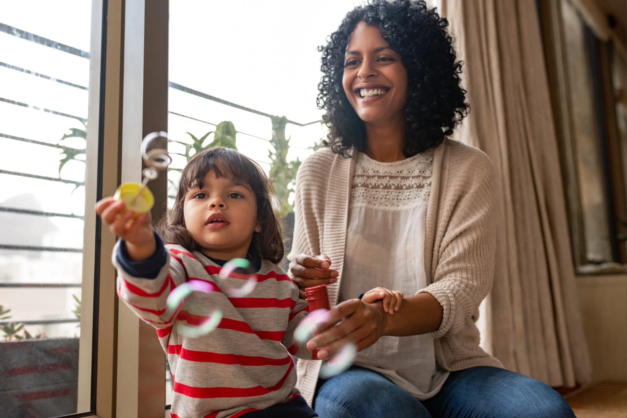A speech therapist and a young girl sitting by a window, playing with bubble wands and bubbles, during in-home speech therapy for children in Wendell, NC.