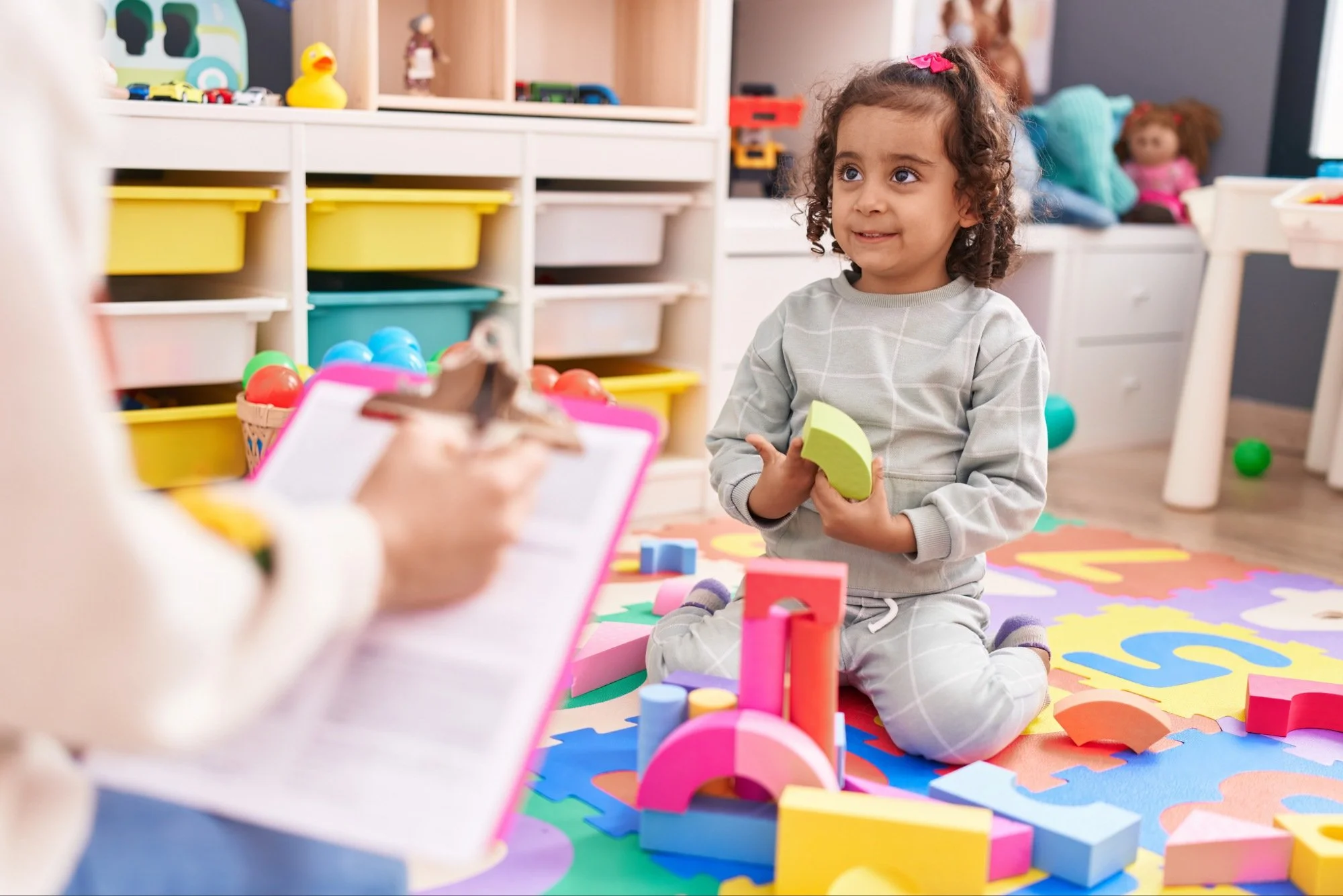 A young girl sitting cross-legged on a colorful foam play mat, holding a green foam block, working with a speech therapist during speech therapy for kids at her daycare center in Zebulon, NC.