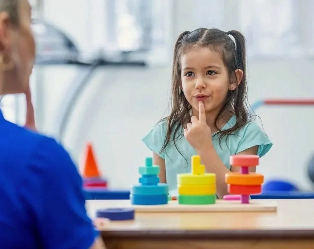 A young girl with dark hair in pigtails working on speech sounds during speech therapy for kids in Zebulon, NC. In front of her are colorful stacking toy rings on a wooden board.