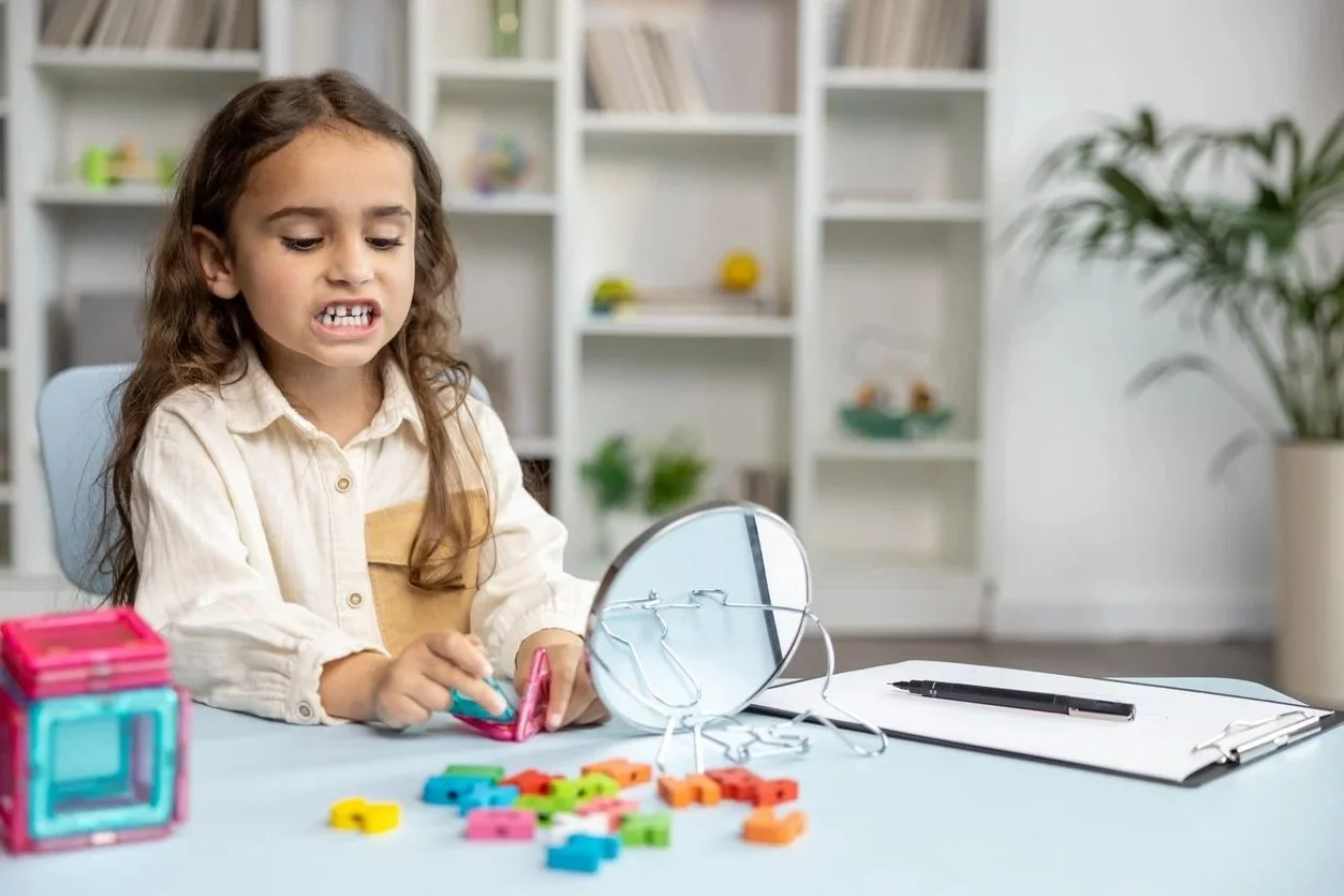 A young girl with long brown hair sitting at a desk with colorful alphabet toys and a mirror, working on making speech sounds during speech therapy for kids in Zebulon, NC.