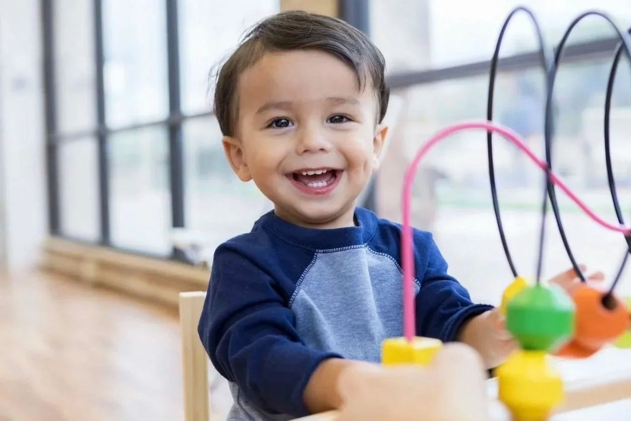 Young boy smiling and playing with a bead maze toy at a table near large windows during speech therapy for children in Zebulon, NC