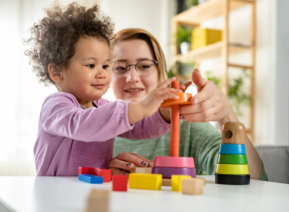 A young girl and a speech therapist working on language skills during speech therapy, playing with colorful stacking ring toys at a table.