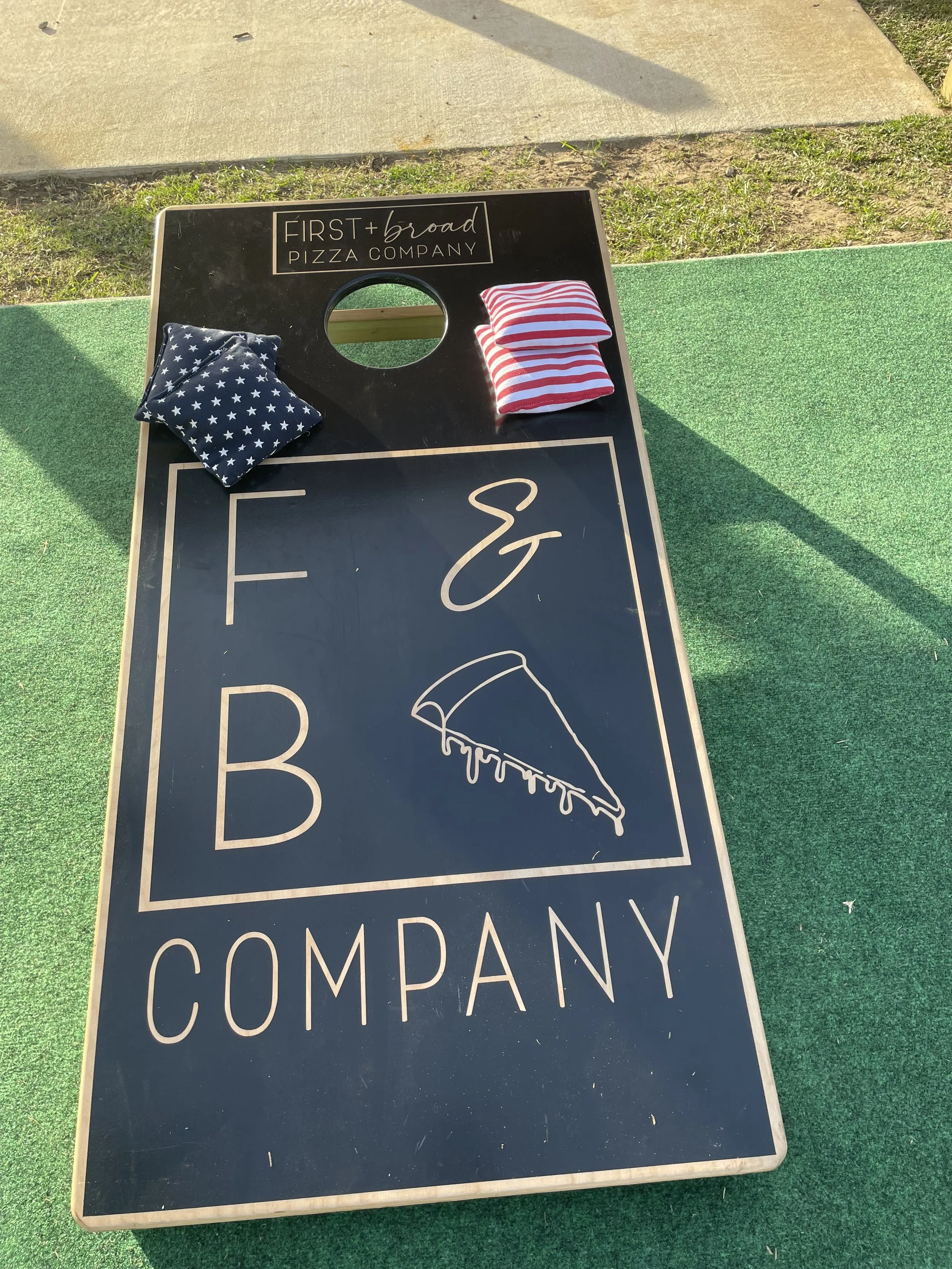 Black cornhole game board for First & Broad Pizza Company with a slice of pizza illustration. Cornhole bags in blue with white stars and red with white stripes are placed on the board.