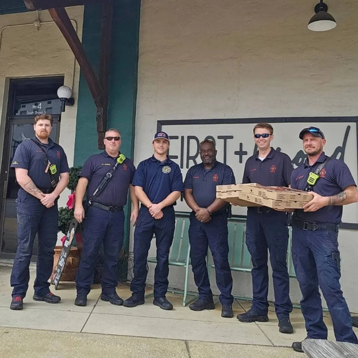 Group of six firefighters standing outside a restaurant, some holding pizza boxes, smiling for a photo.