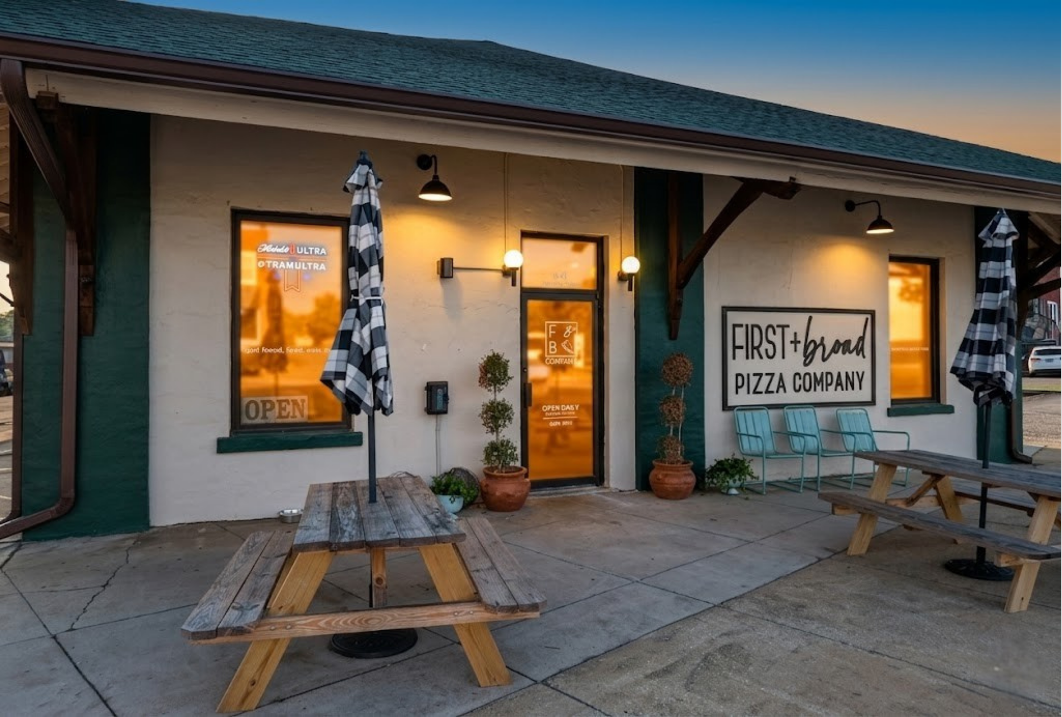 The exterior of First + Broad Pizza Company during sunset, featuring outdoor seating with picnic tables, potted plants, umbrellas, and illuminated windows and signs.