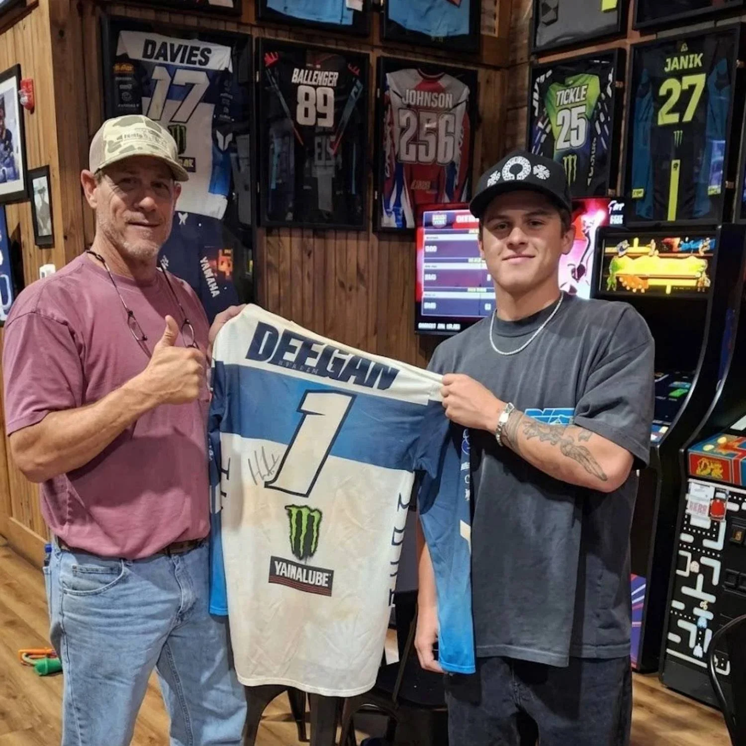 Two men standing indoors, holding a race jersey.  The background features framed sports jerseys on wooden walls and arcade machines.
