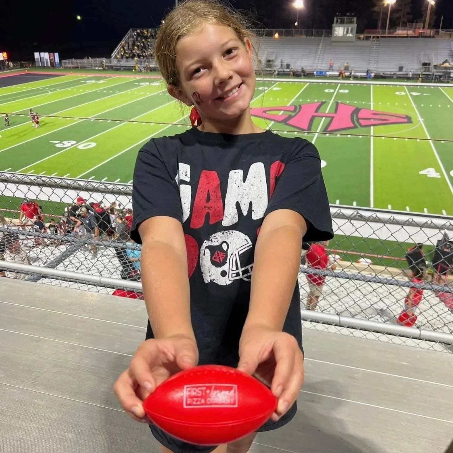 Young girl at a football stadium holding a red football, smiling, with the field and players in the background during nighttime.