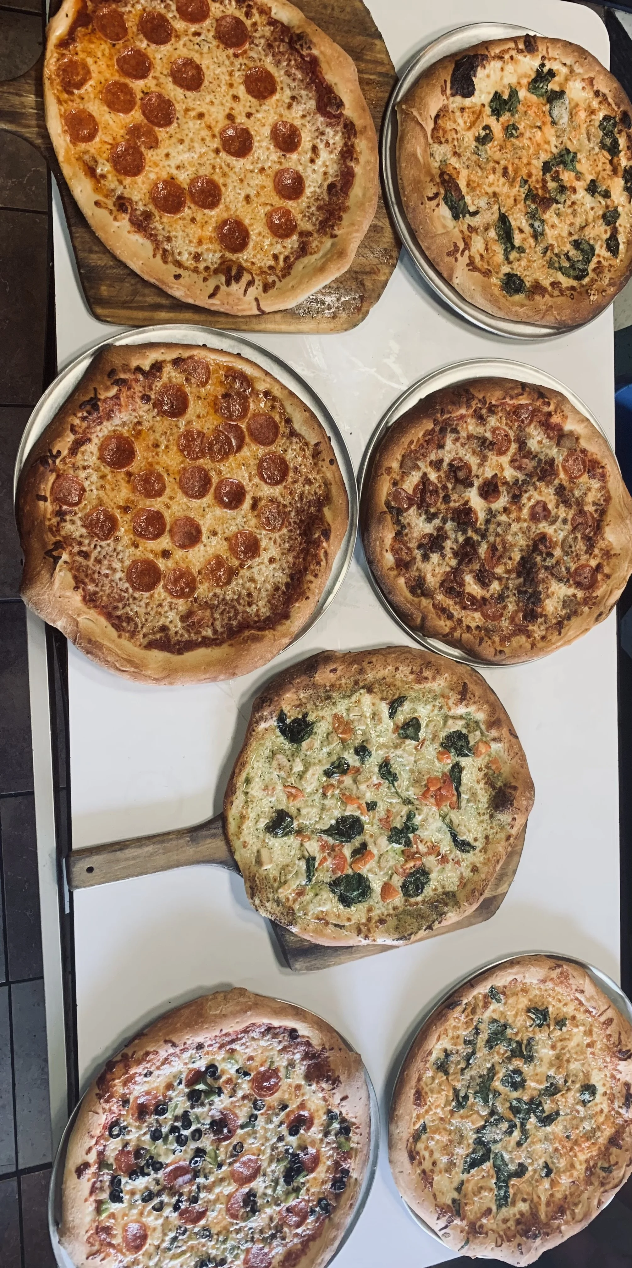 Eight pizzas with various toppings, some with pepperoni, vegetables, and cheese, on a white table, with some on wooden boards.