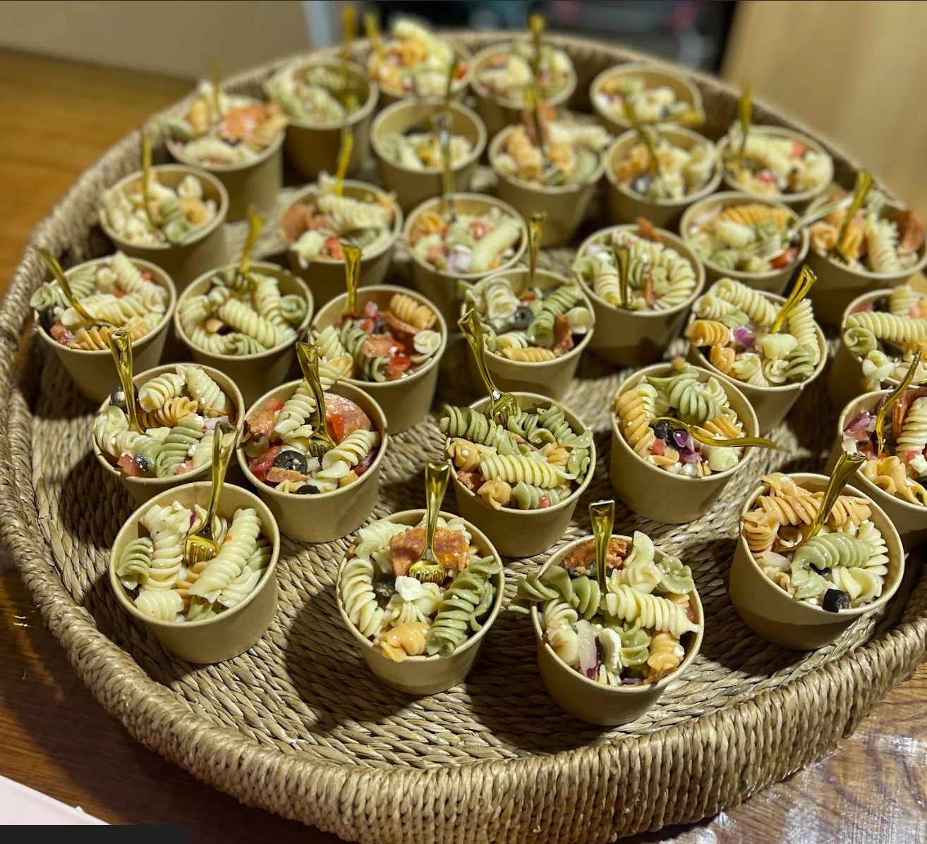 Small cups filled with colorful pasta salad, each with a small serving fork, arranged on a round woven tray.