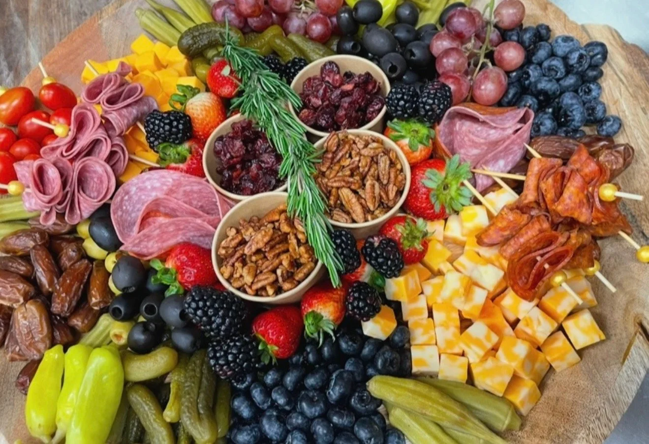 A charcuterie board with cheeses, grapes, strawberries, blackberries, blueberries, pickles, cherry tomatoes, cubed cheese, sliced meats, dried cranberries, nuts, and fresh herbs.