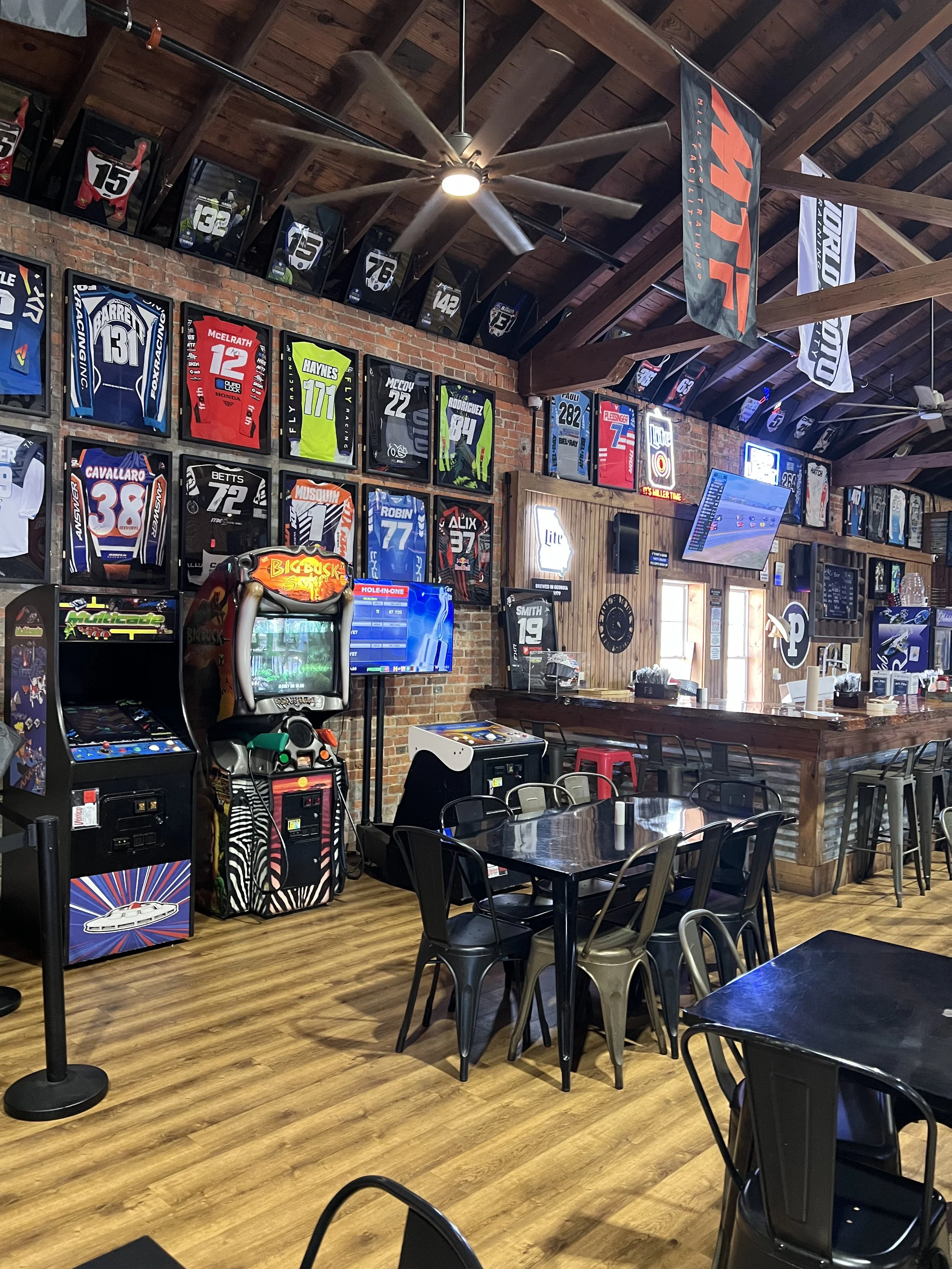 A sports bar with framed jerseys on the brick wall, arcade game machines, a bar with stools, tables and chairs, and multiple flat-screen TVs mounted on the wall.