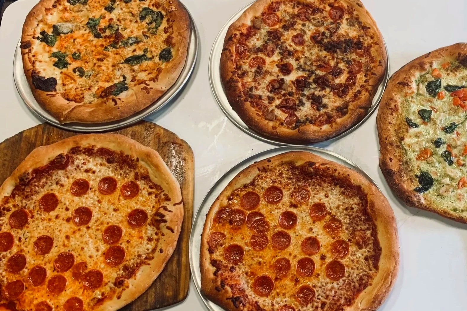 Five pizzas with different toppings on metal trays and a wooden cutting board, including pepperoni, sausage, vegetables, and cheese.