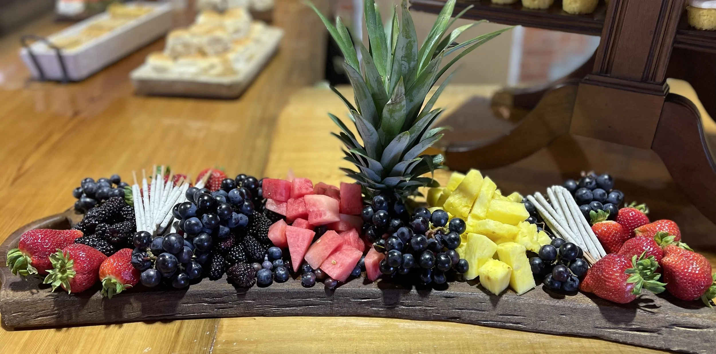 A wooden tray with an array of fresh fruits including strawberries, blackberries, blueberries, watermelon, yellow pineapple, and cubed watermelon in the center, with a pineapple on top for decoration.
