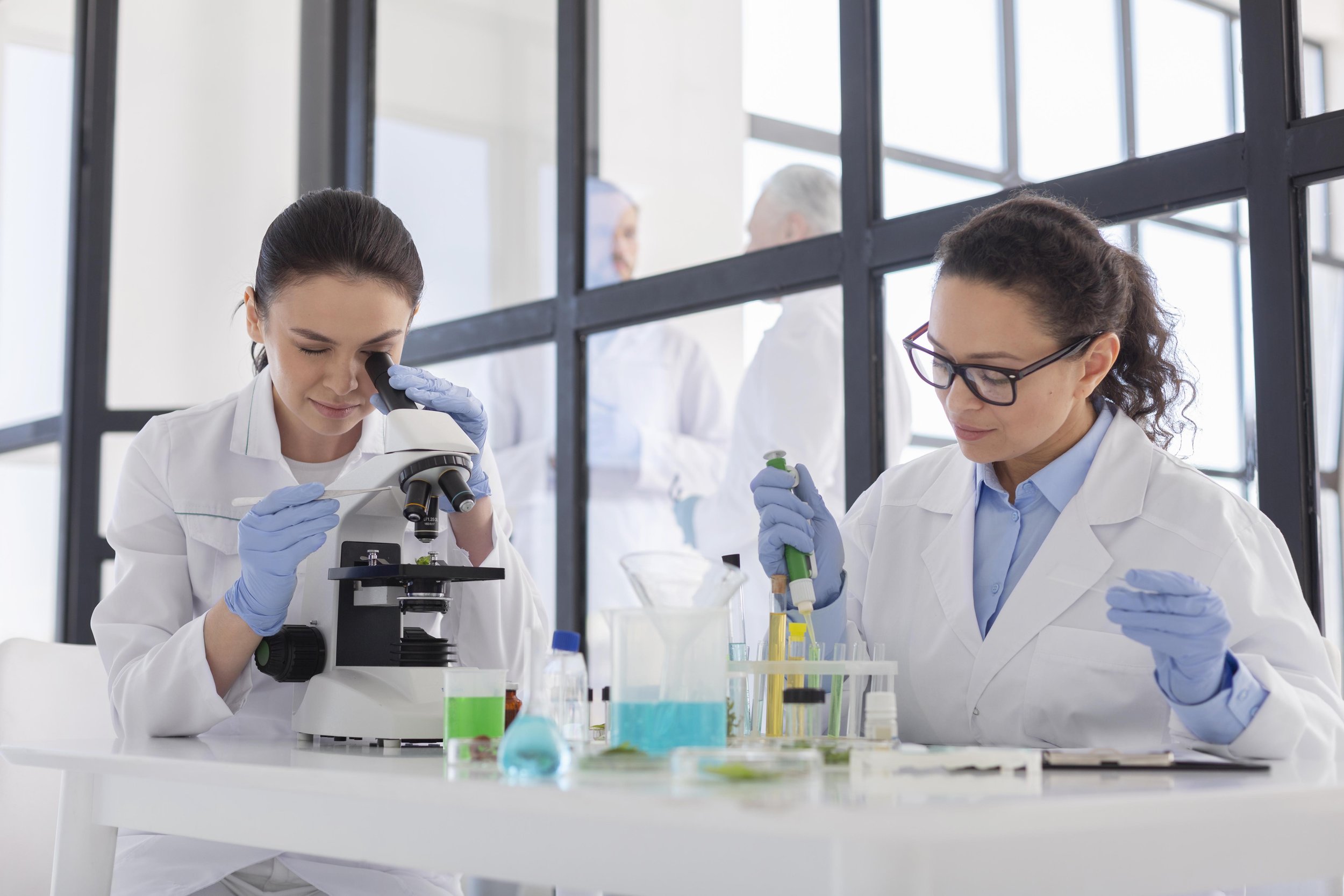 Two female scientists working in a laboratory with scientific equipment, including a microscope, test tubes, and chemicals.