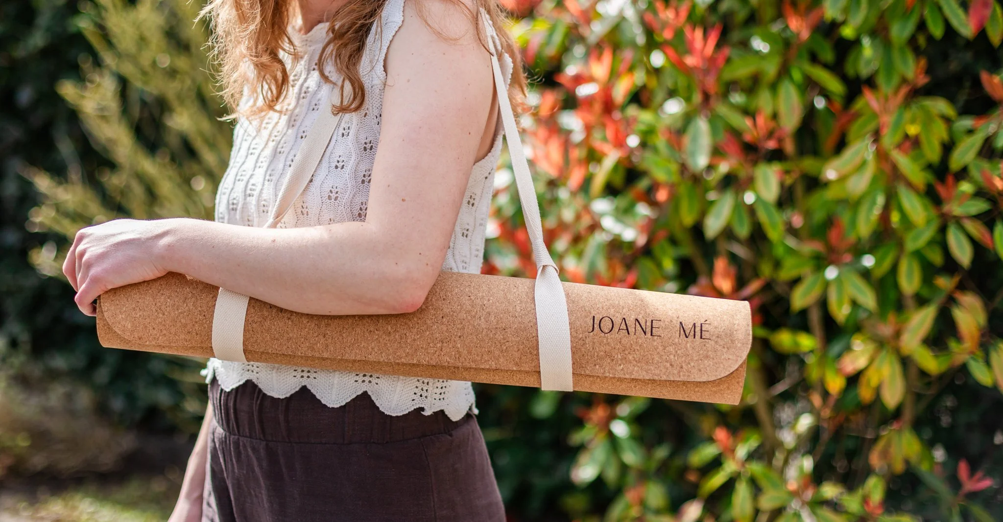 Een vrouw met een witte kanten top en een paarse rok houdt een kartonnen tas met de naam 'JOANE MÉ' in een buitenomgeving met groene en oranje bladeren.