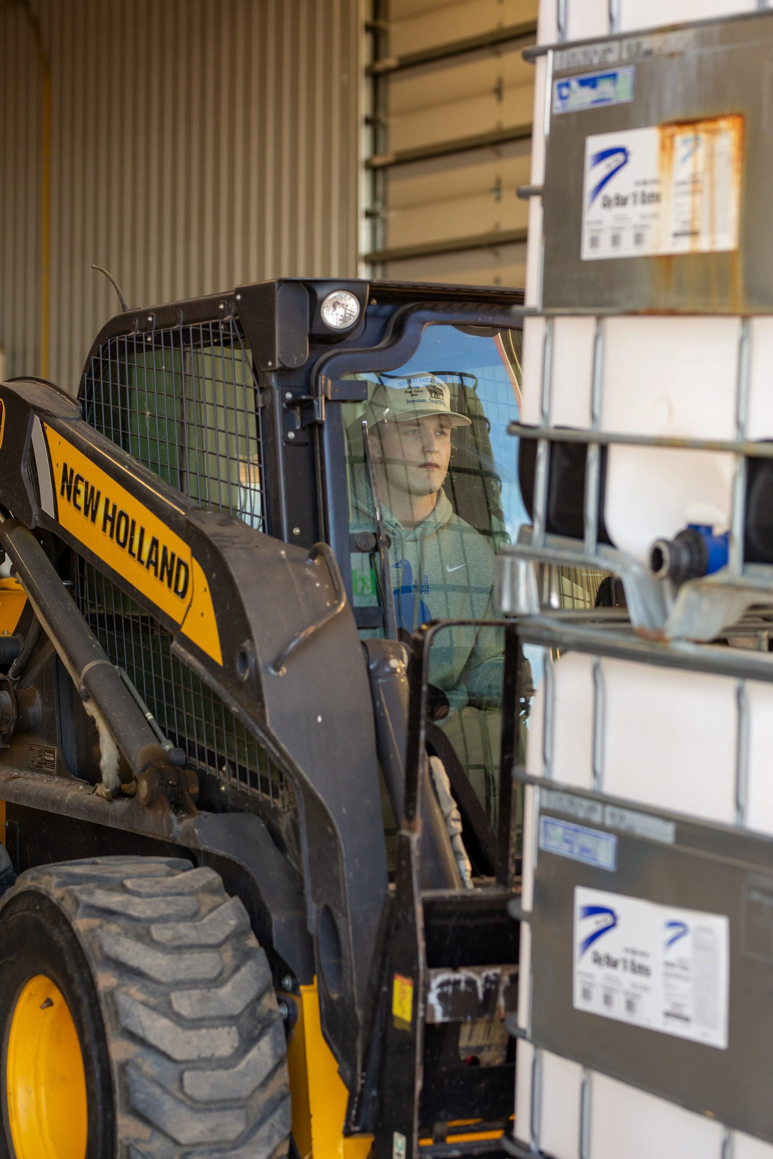 A young man wearing a baseball cap and hoodie sitting inside a New Holland skid steer loader in a warehouse or garage.