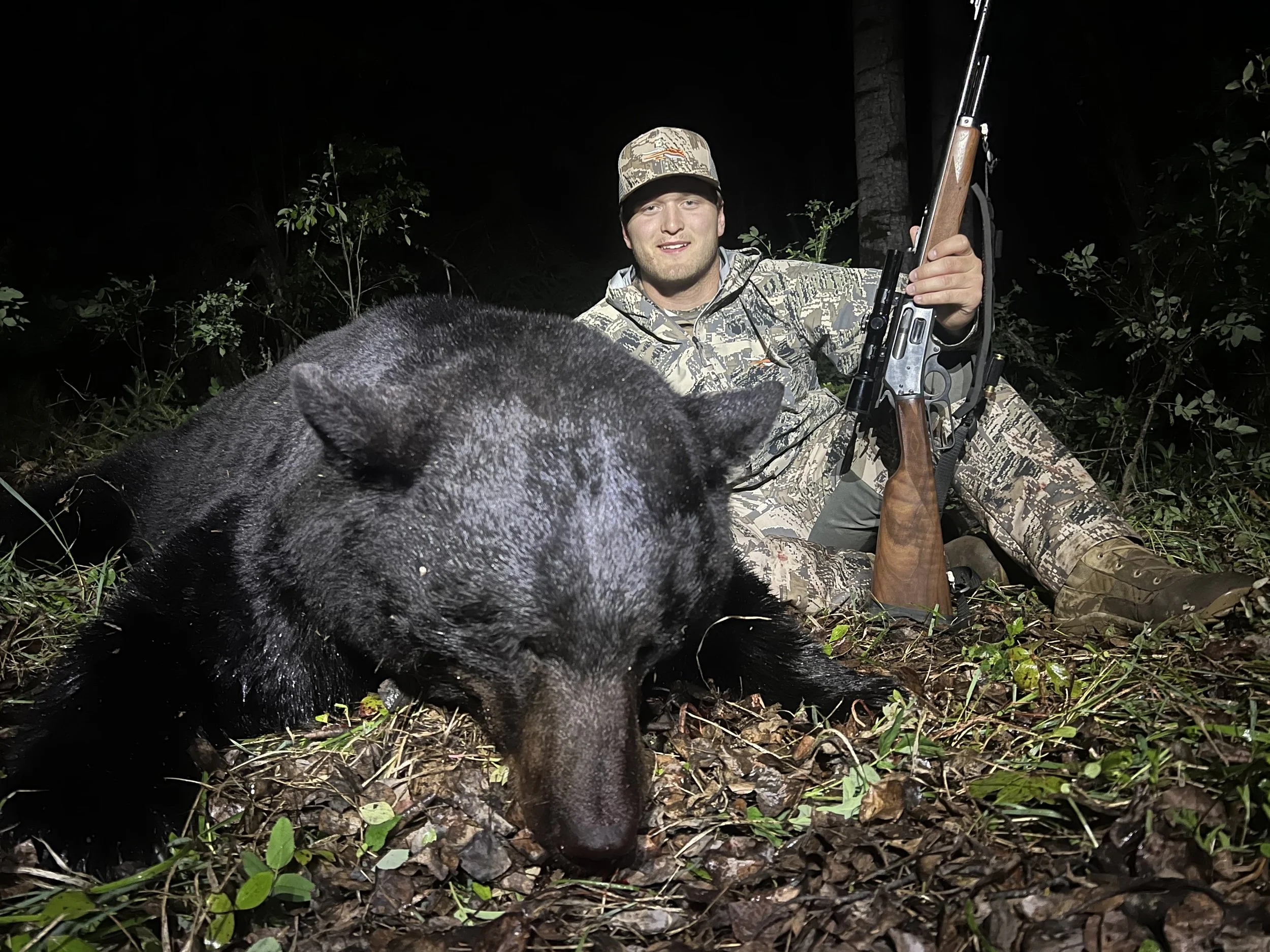 A man in camouflage attire sitting in the woods at night, holding a rifle, with a black bear lying on the ground in front of him.