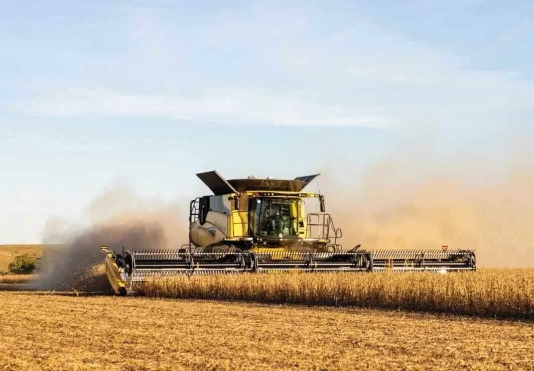 A large combine harvester machine working in a crop field during daytime, harvesting grains with dust clouds in the background.